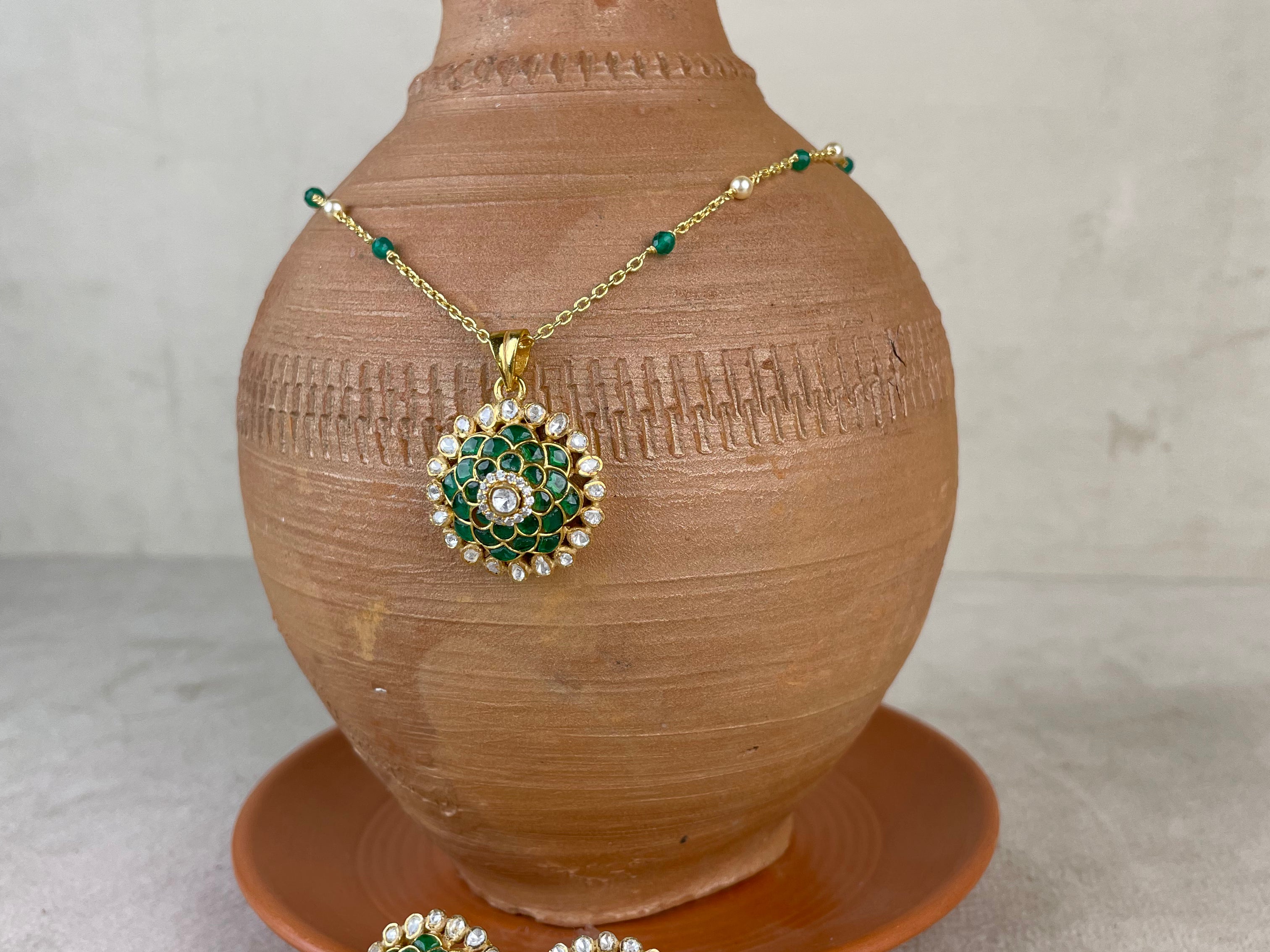 Gold and green jewelry set displayed on a terracotta vase, featuring a necklace with a round pendant and matching earrings with a white price tag attached.