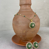 Gold-tone jewelry set with a pendant and matching earrings, featuring green stone floral motifs and white stone accents, displayed on a terracotta vase and saucer against a neutral background.