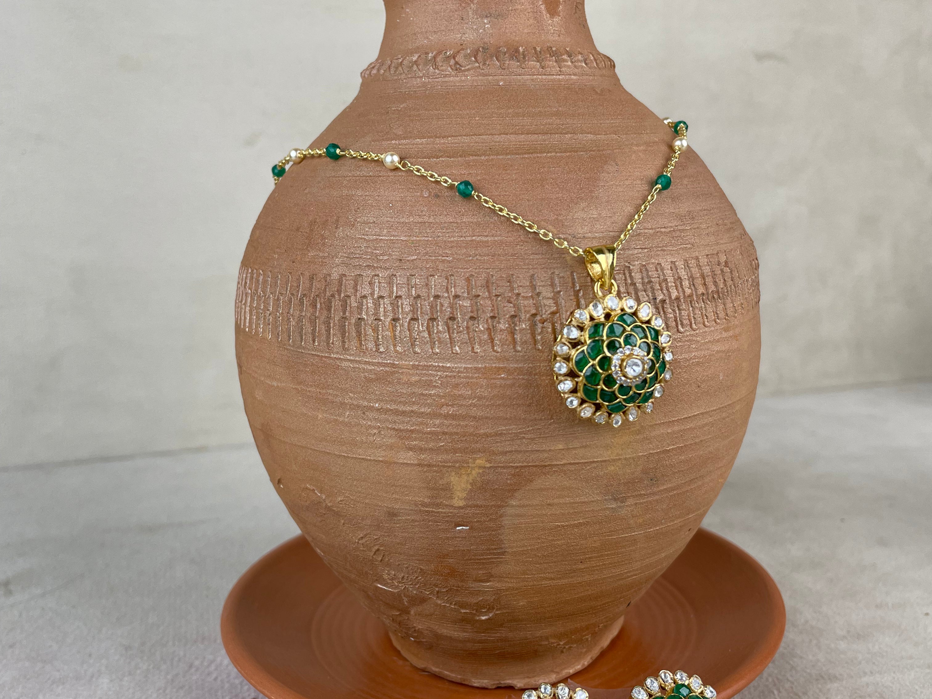 Gold-tone jewelry set with a pendant and matching earrings, featuring green stone floral motifs and white stone accents, displayed on a terracotta vase and saucer against a neutral background.