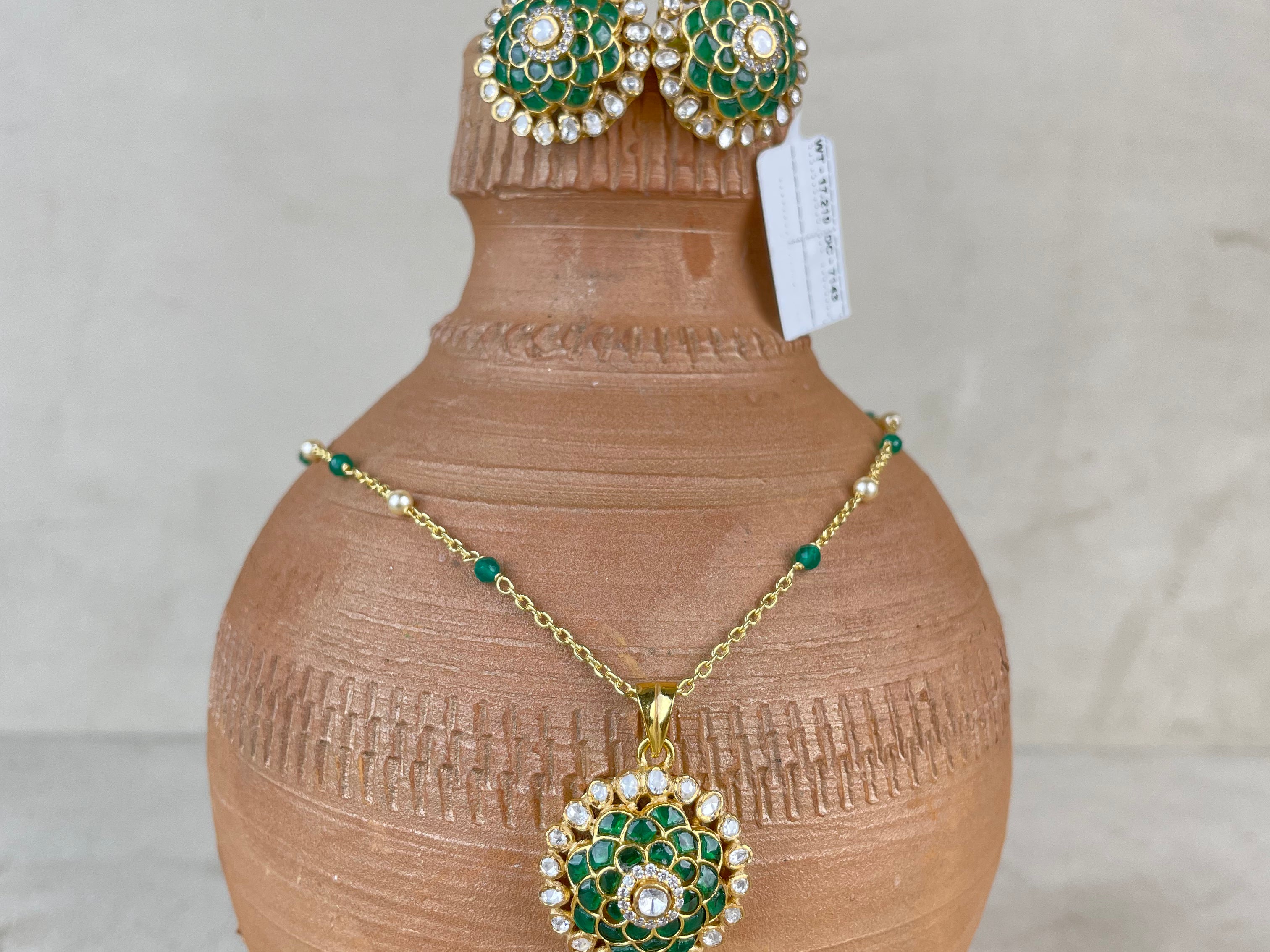 A gold jewelry set featuring a necklace and matching earrings with green and white enamel detailing, displayed on a terracotta vase against a neutral background.