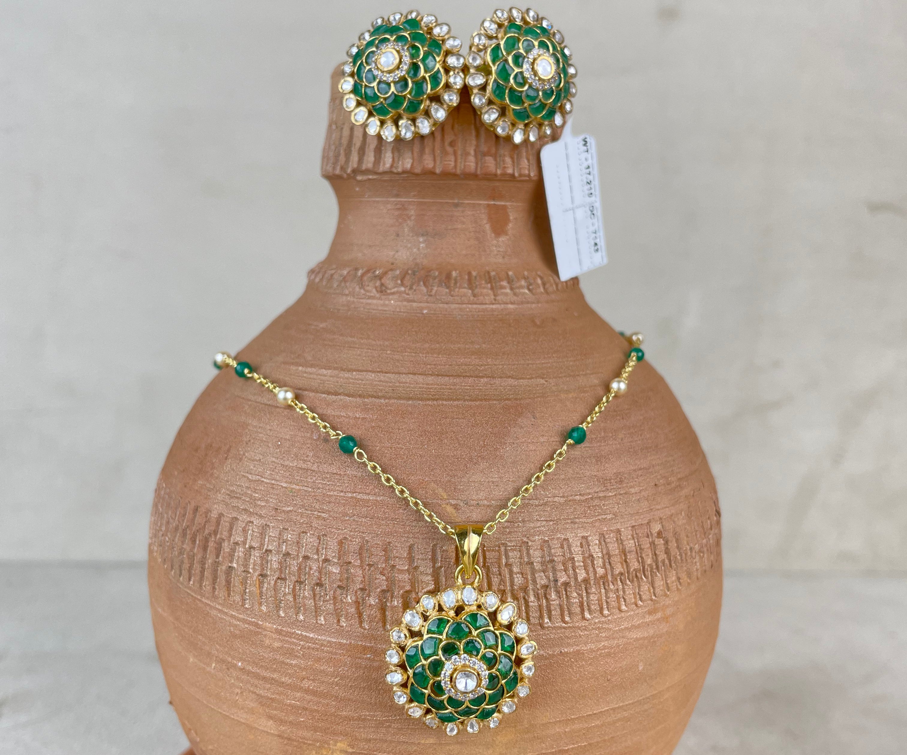 A gold jewelry set featuring a necklace and matching earrings with green and white enamel detailing, displayed on a terracotta vase against a neutral background.