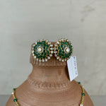 A pair of ornate green and gold floral stud earrings with pearl-like accents, displayed on a terracotta jewelry bust alongside a matching green and gold necklace against a neutral light gray background.