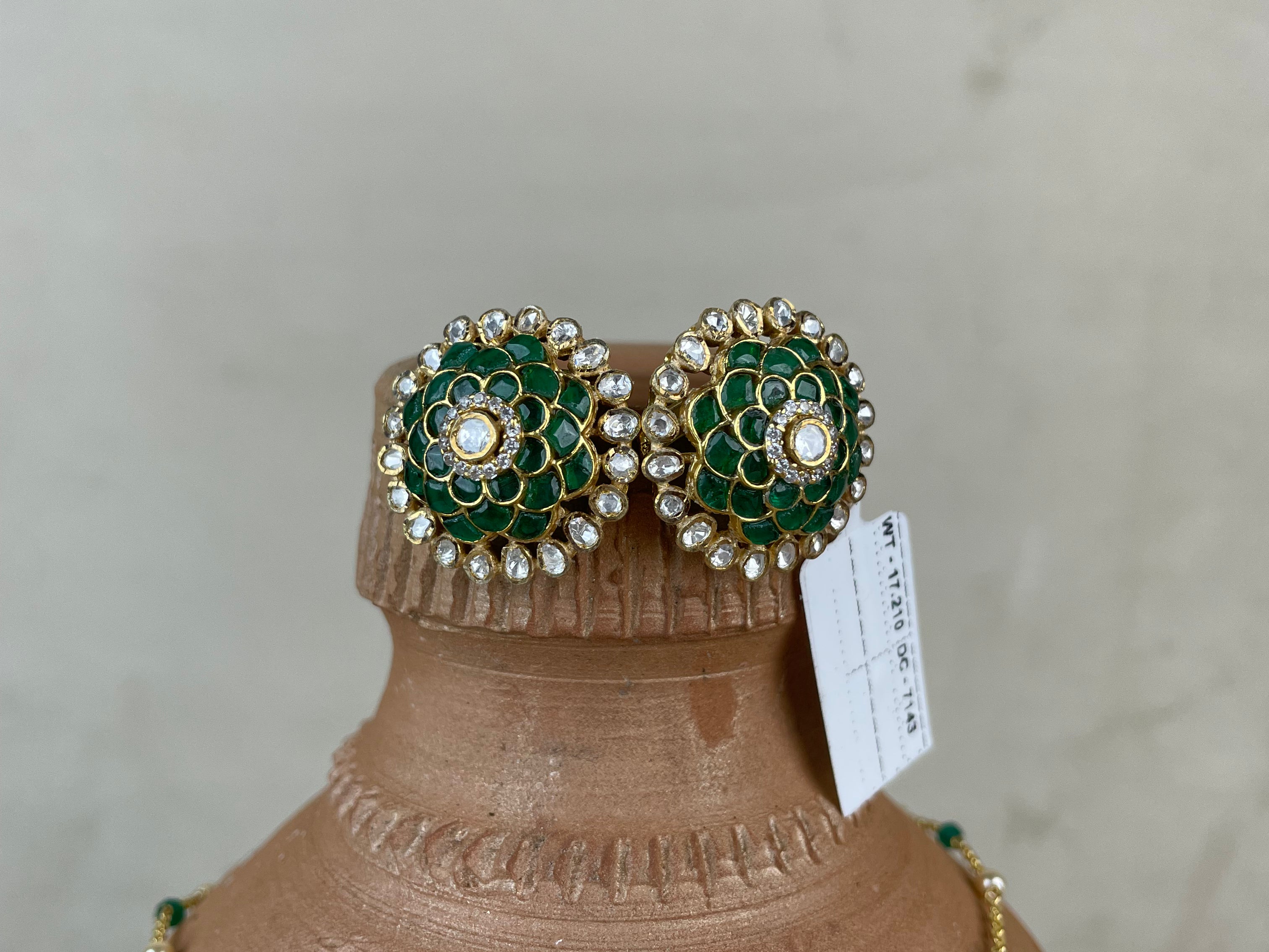 A pair of ornate green and gold floral stud earrings with pearl-like accents, displayed on a terracotta jewelry bust alongside a matching green and gold necklace against a neutral light gray background.
