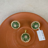 A jewelry set featuring a gold pendant and matching earrings with green enamel floral detailing and white stones, displayed on a terracotta-colored ceramic plate against a neutral background.