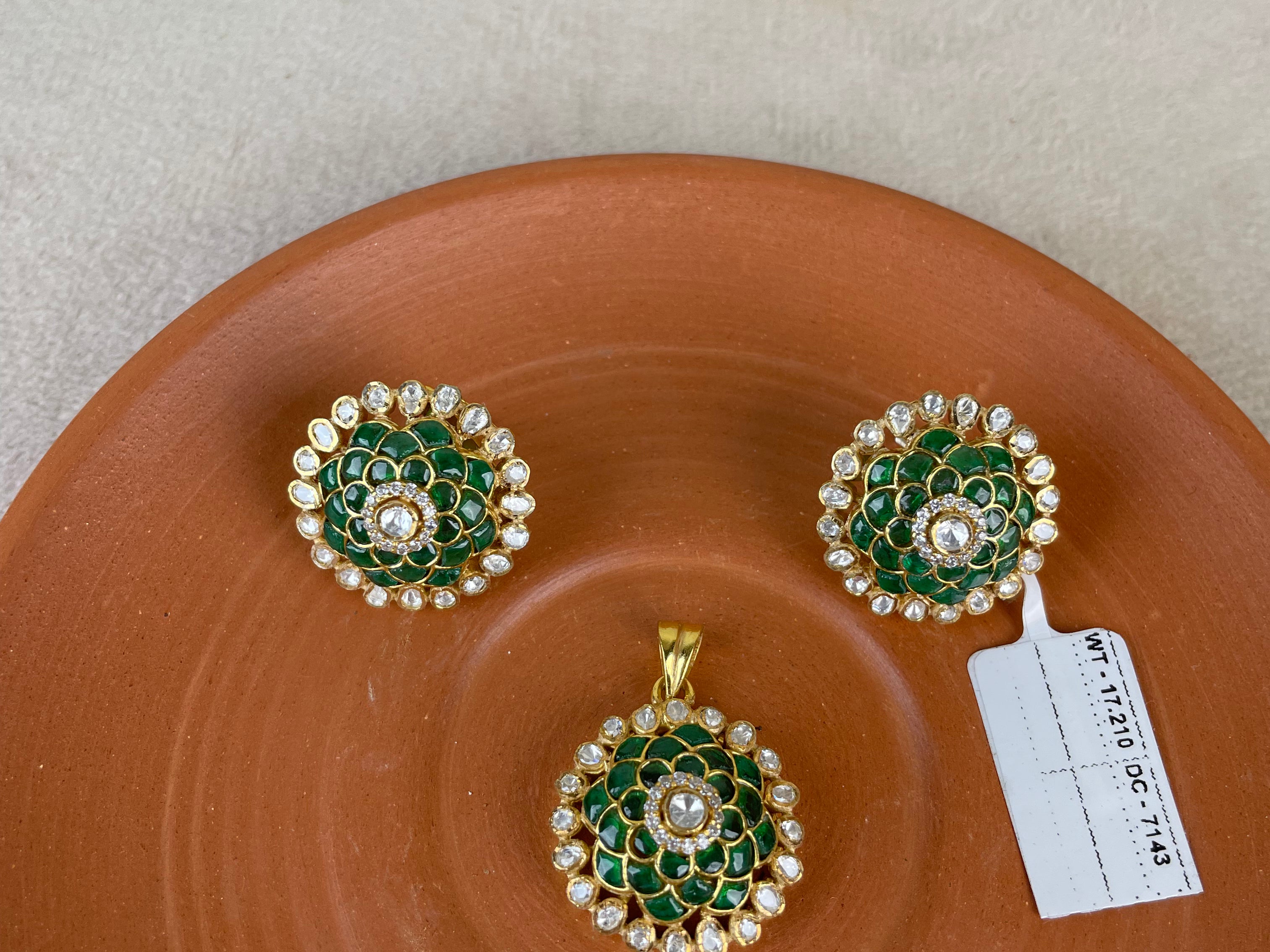 A jewelry set featuring a gold pendant and matching earrings with green enamel floral detailing and white stones, displayed on a terracotta-colored ceramic plate against a neutral background.
