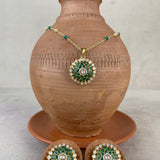 Gold and green jewelry set displayed on and around a terracotta vase, featuring a round pendant necklace and two matching earrings with green enamel, gold detailing, and clear stones.
