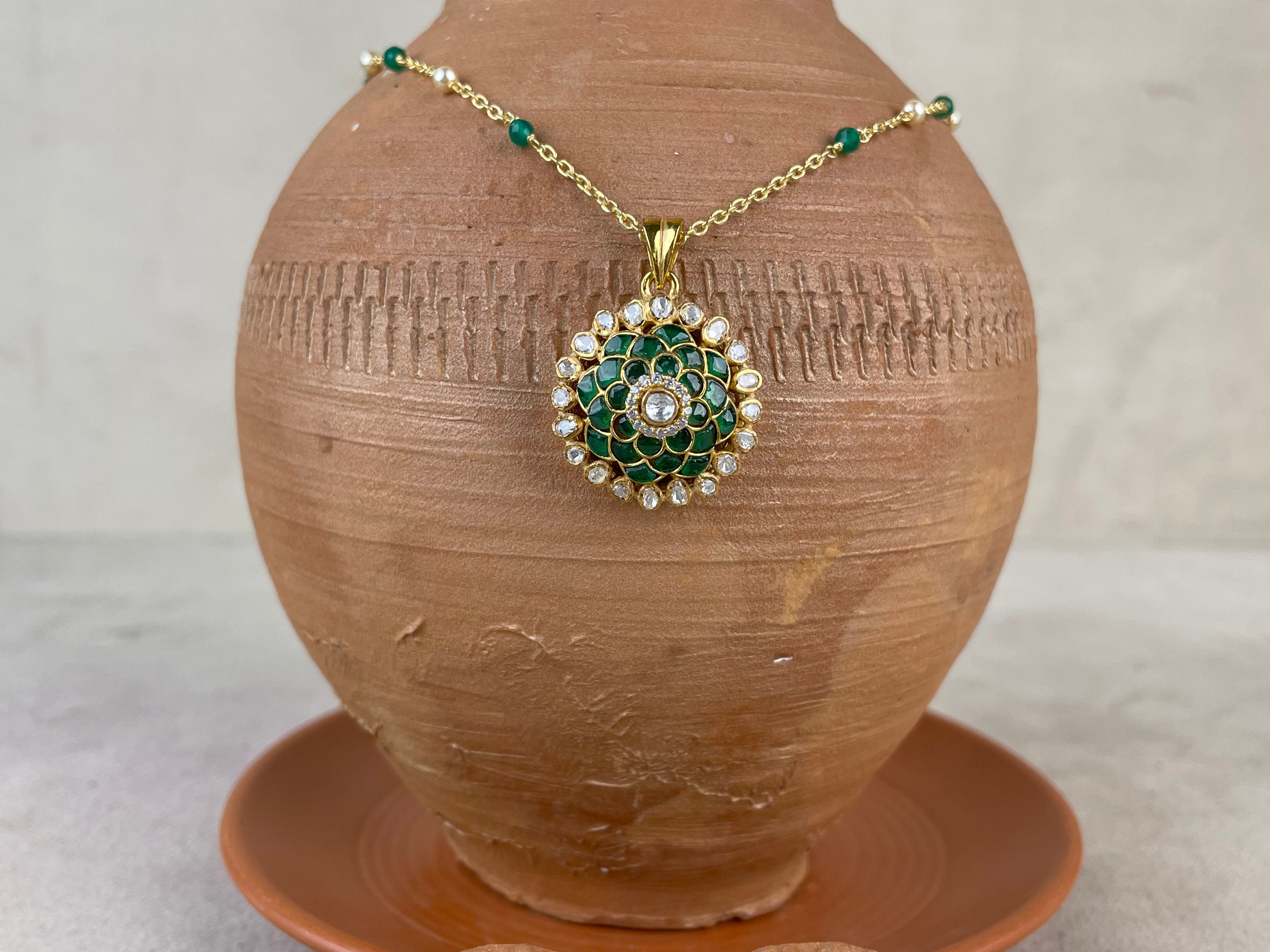 Gold and green jewelry set displayed on and around a terracotta vase, featuring a round pendant necklace and two matching earrings with green enamel, gold detailing, and clear stones.