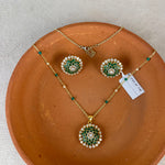 Gold jewelry set with a pendant and matching earrings featuring green stone and white pearl floral motifs, displayed on a terracotta plate over a light beige fabric background.