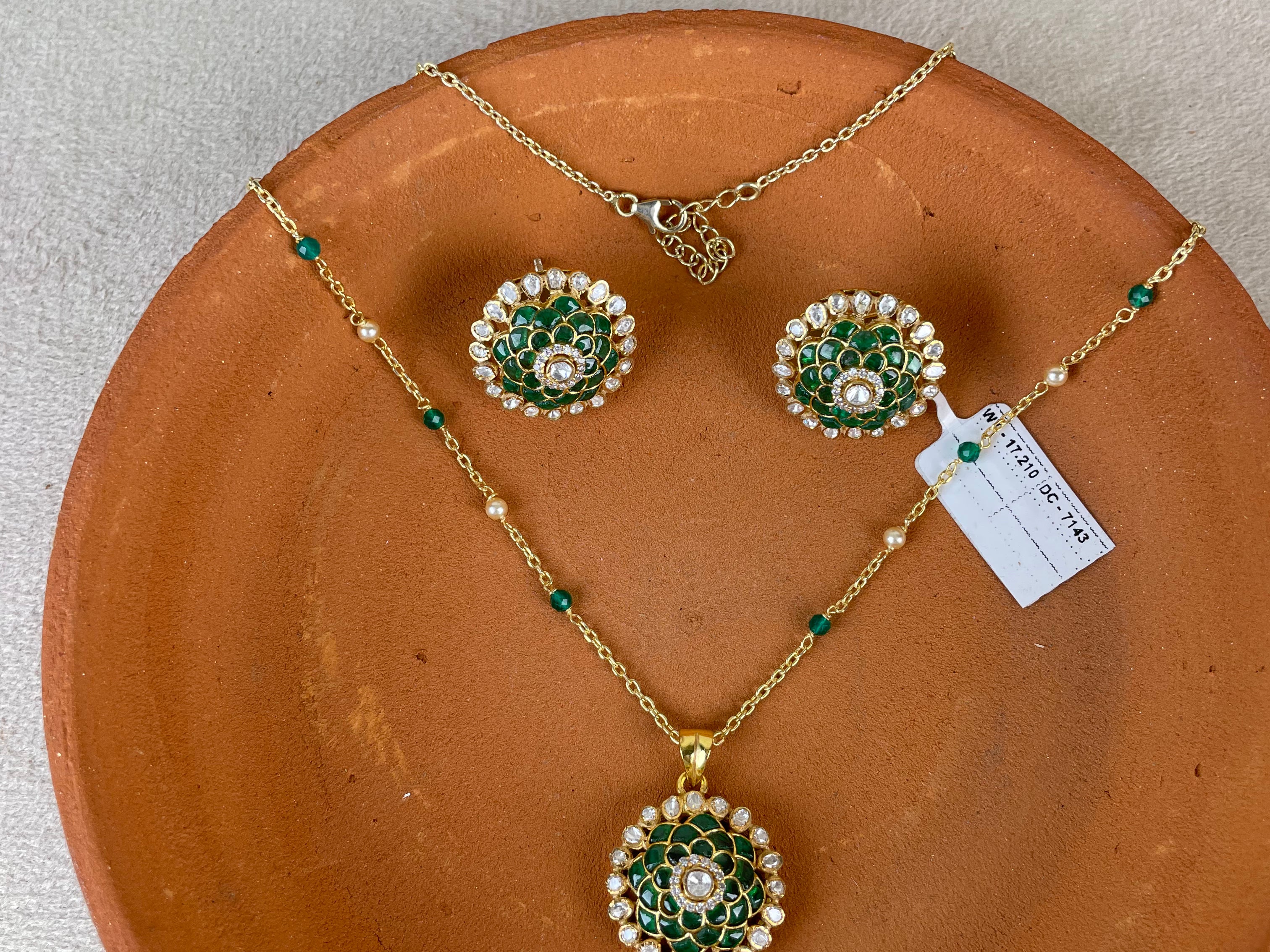 Gold jewelry set with a pendant and matching earrings featuring green stone and white pearl floral motifs, displayed on a terracotta plate over a light beige fabric background.
