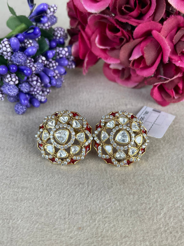A pair of ornate gold-toned earrings with clear stones and red enamel accents, displayed on a light beige textured surface with decorative pink and purple flowers in the background.