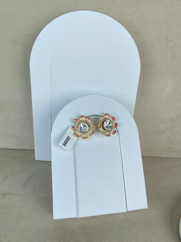 A pair of gold-toned floral stud earrings with clear central stones and pink/red accents, displayed on a white arched jewelry stand against a beige background.