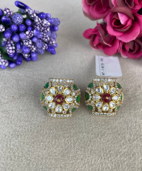 A pair of ornate gold-toned Indian Kundan earrings decorated with green enamel, clear crystals, and a central red stone, displayed on a textured beige surface with decorative flowers in the background.