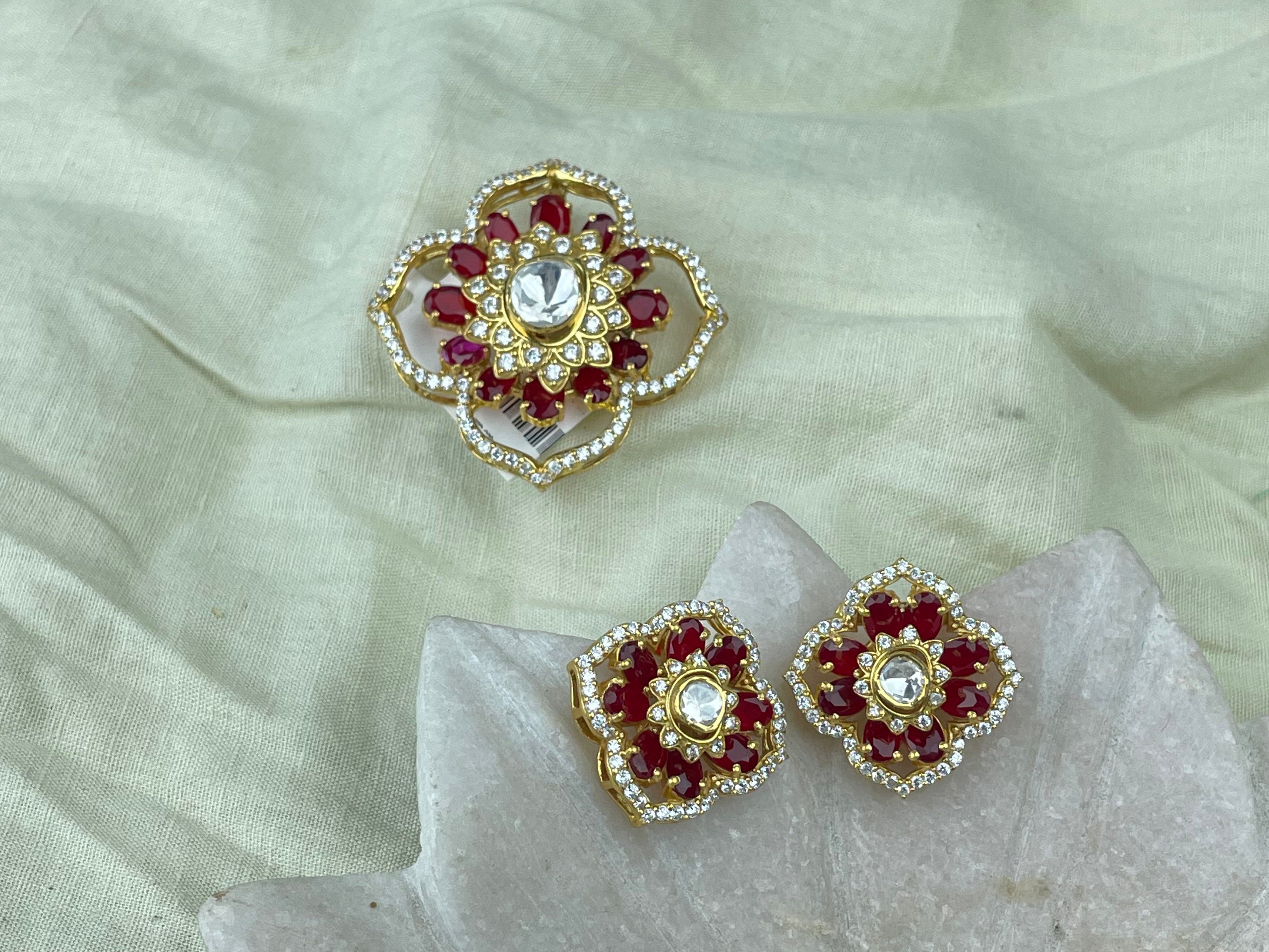 Pendant Set with matching a pair of gold-toned floral stud earrings with red stones and a central clear stone, displayed on a pale fabric surface with a matching larger decorative piece above.