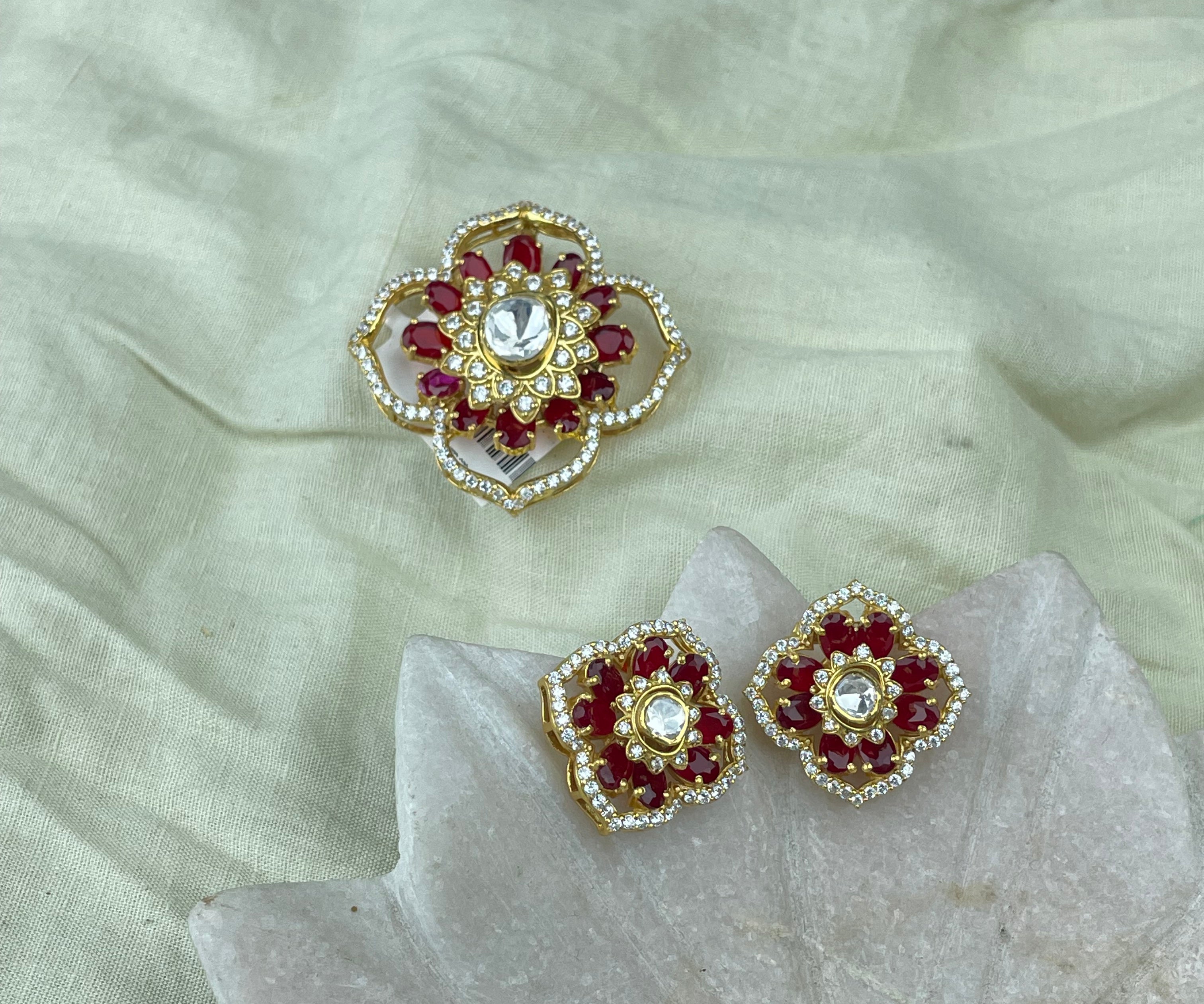 Pendant Set with matching a pair of gold-toned floral stud earrings with red stones and a central clear stone, displayed on a pale fabric surface with a matching larger decorative piece above.