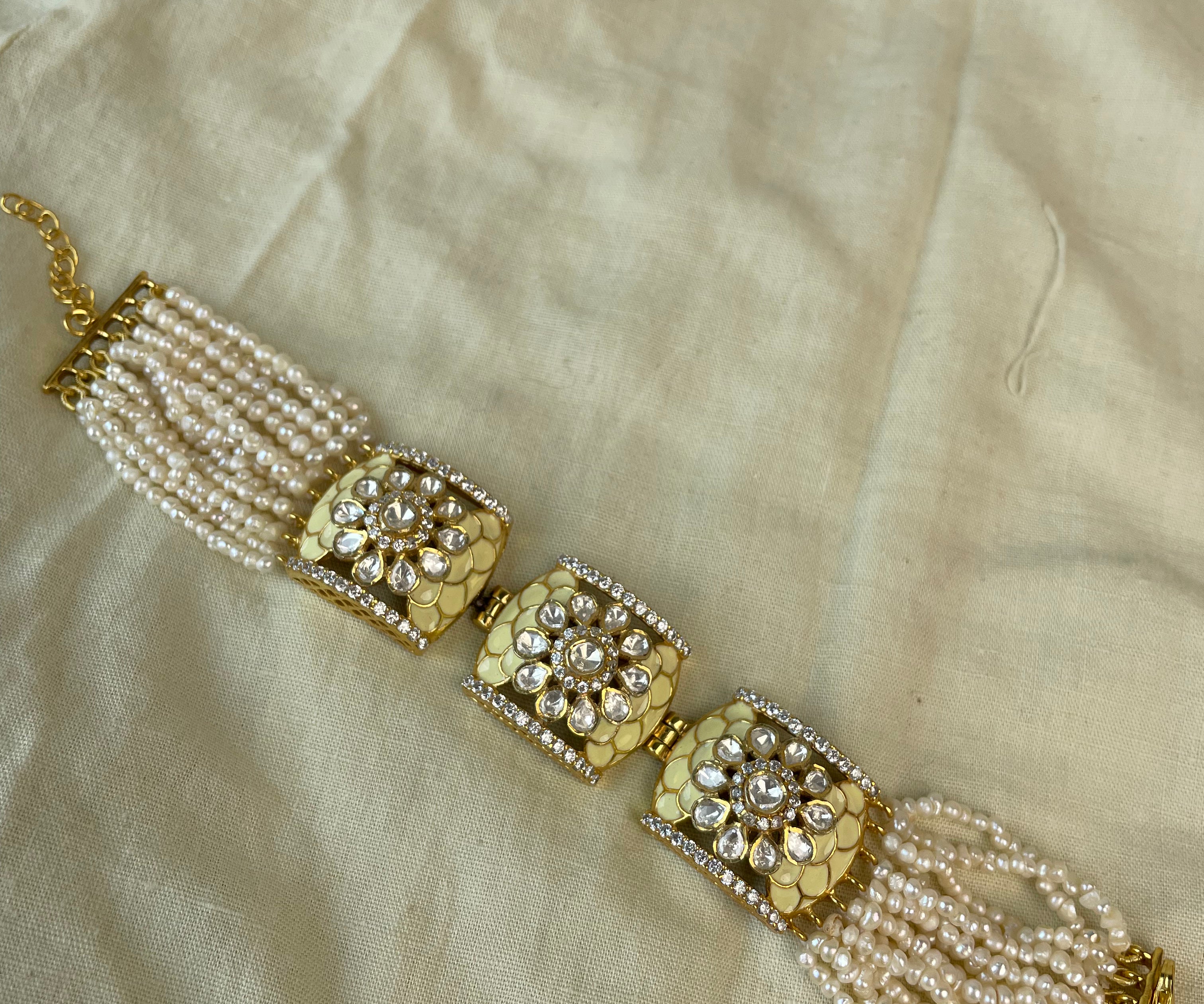 A traditional Indian gold-plated bracelet with white pearl strands and intricate silver and gold floral embellishments, laid flat on a beige fabric background.
