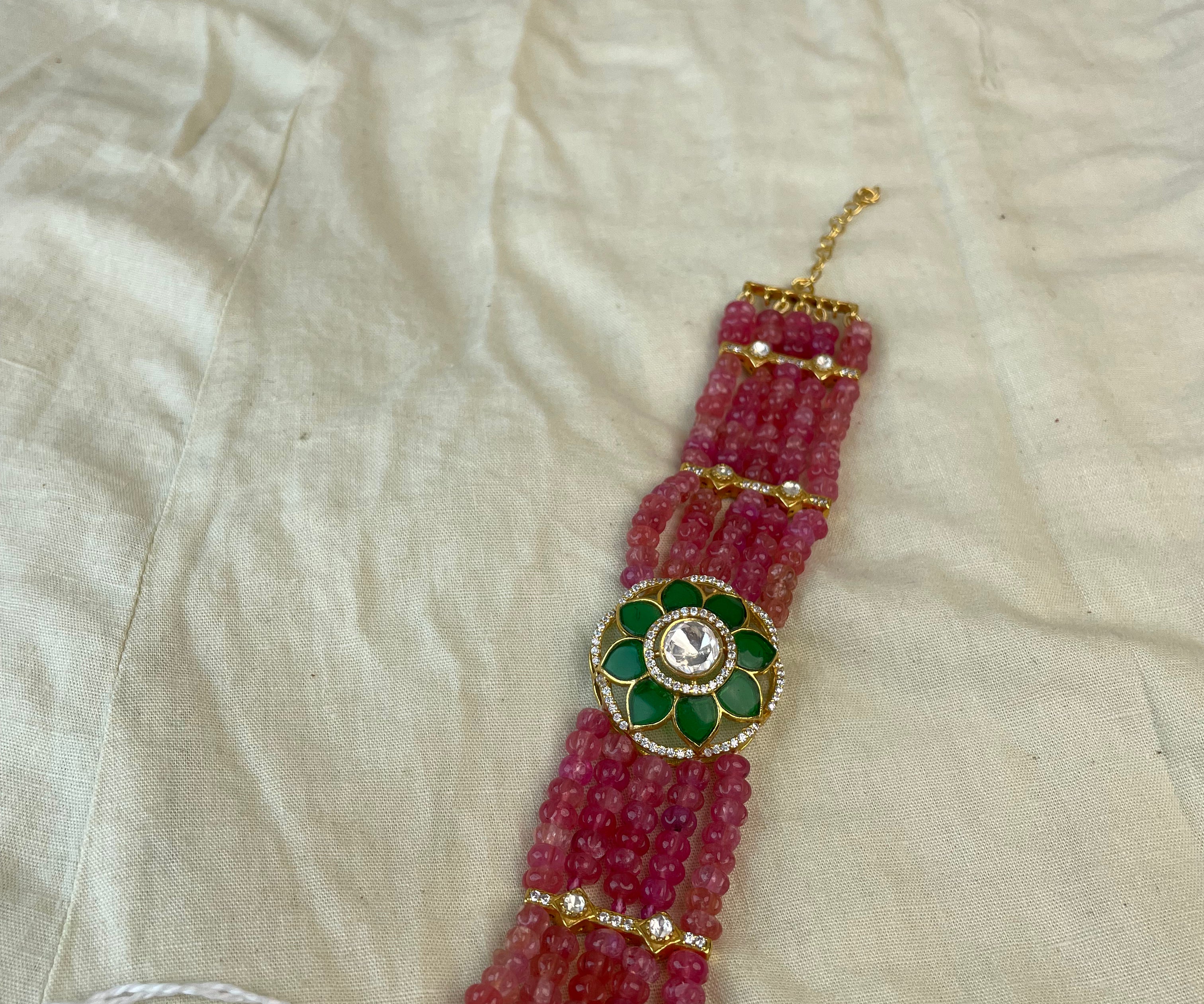 A beaded wristband or bracelet featuring multiple strands of pinkish-red beads, a central green and gold floral medallion with a white stone, and small gold spacer beads, laid on a cream-colored fabric background.