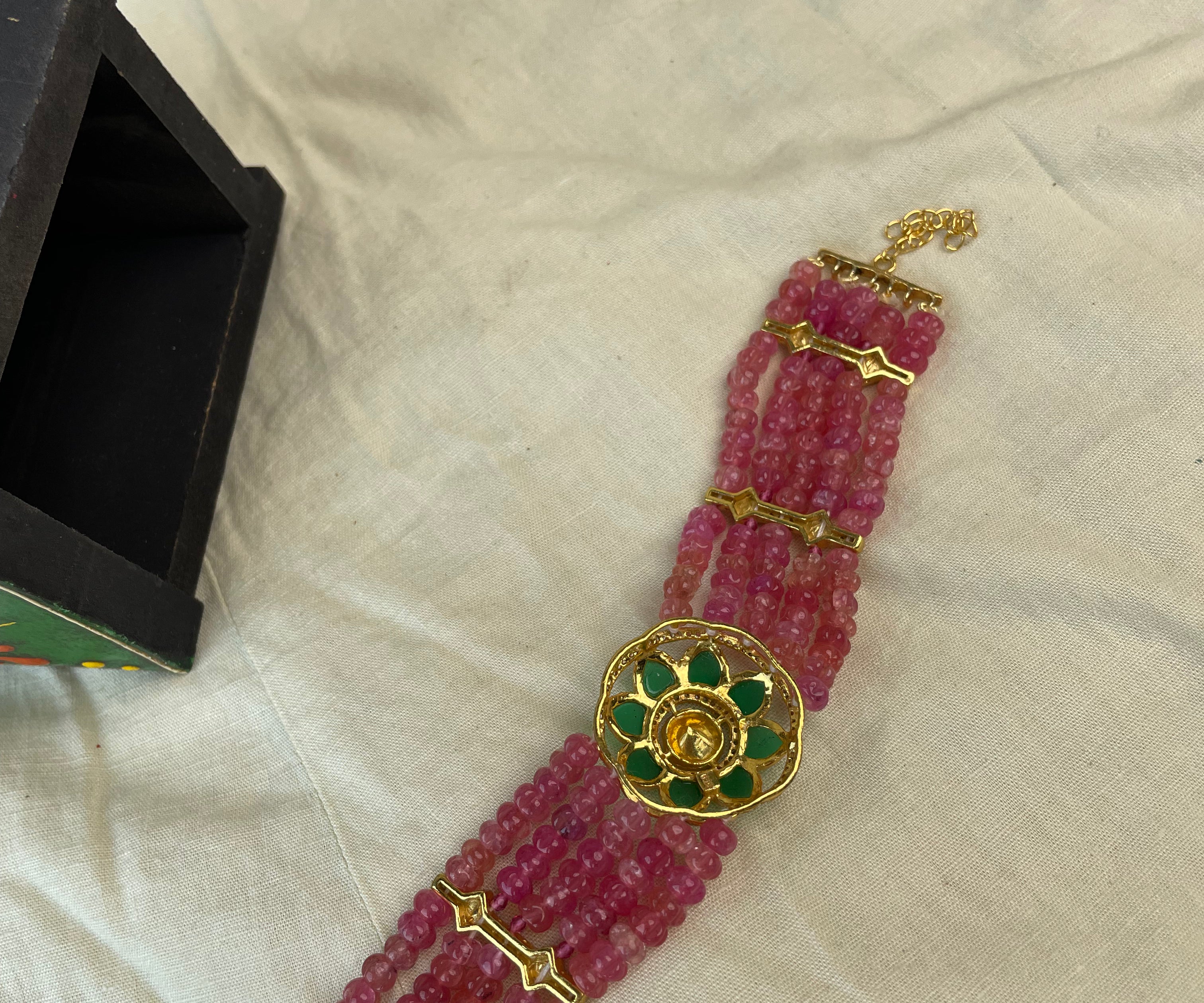 A traditional Indian multi-strand beaded bracelet made of pinkish-red glass beads with gold-plated metal accents and a central round gold and green stone centerpiece, displayed on a beige fabric surface next to a black box.