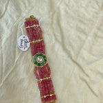 A handmade beaded bracelet made of pink glass beads with gold-toned metal accents and a central green and gold floral motif with a pearl-like center, laid on a cream-colored fabric background with a handwritten tag reading '1500 beads'.