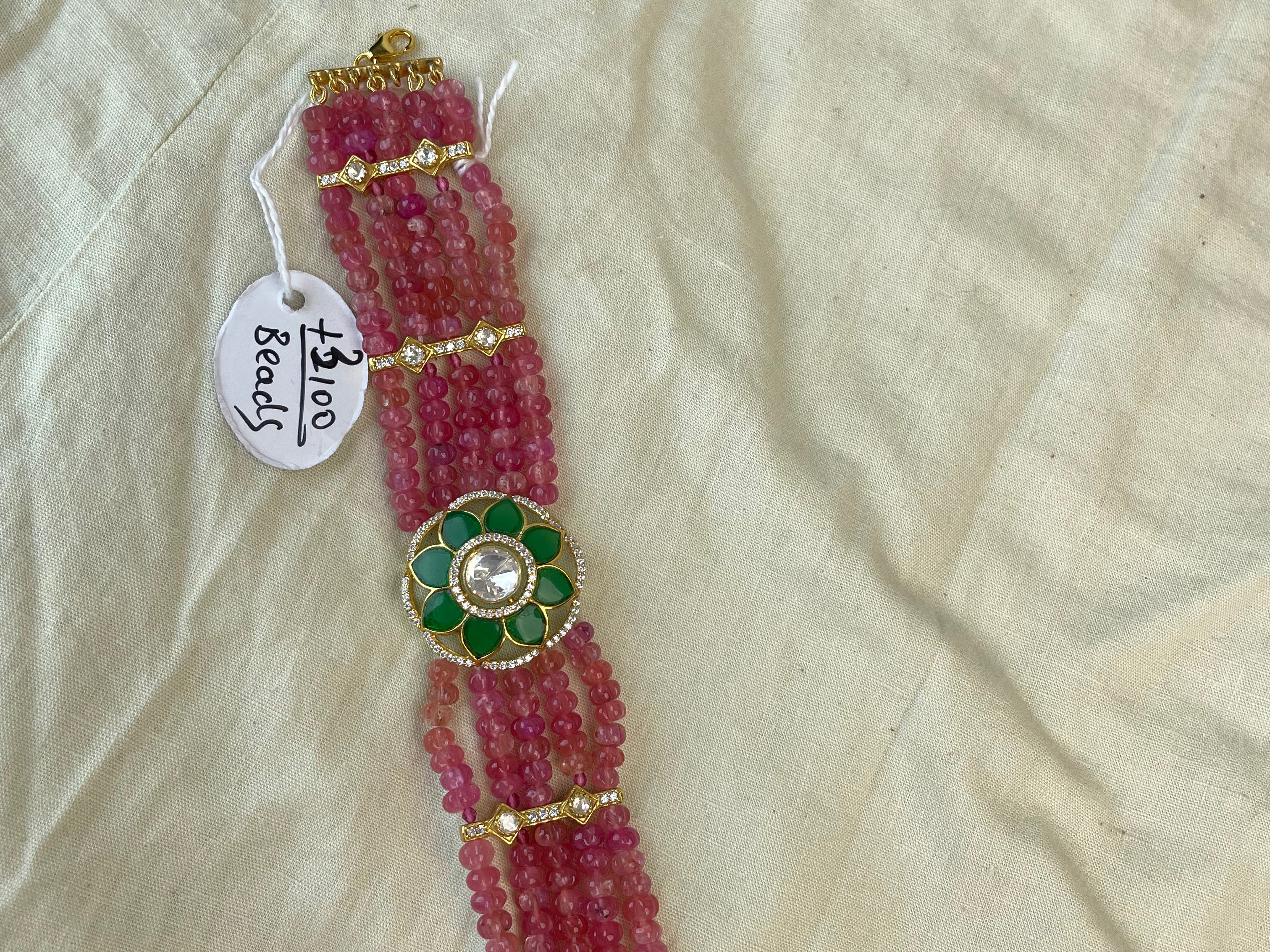 A handmade beaded bracelet made of pink glass beads with gold-toned metal accents and a central green and gold floral motif with a pearl-like center, laid on a cream-colored fabric background with a handwritten tag reading '1500 beads'.