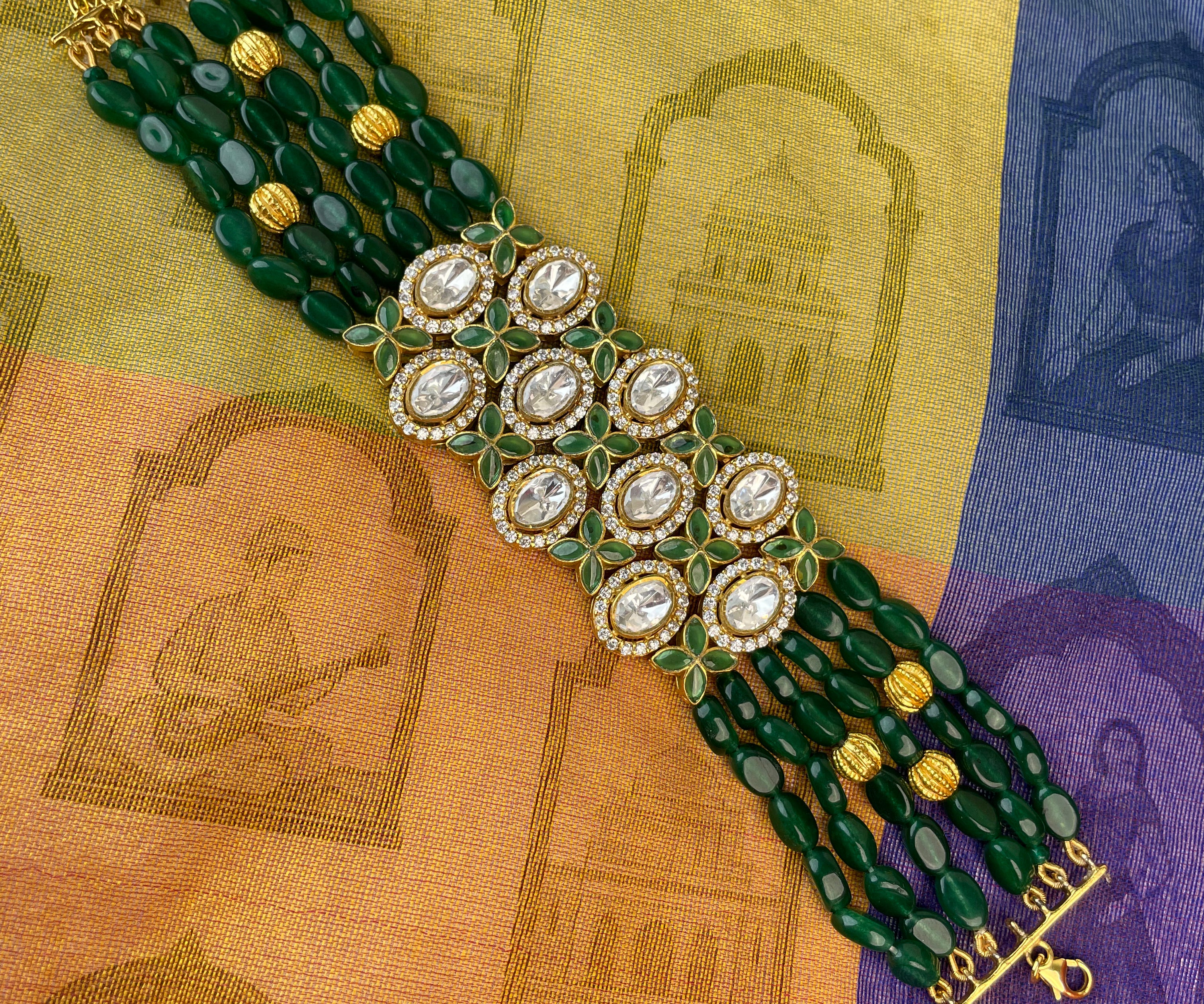 A multi-strand beaded bracelet featuring dark green beads, gold-toned metal details, and a central cluster of round white stones surrounded by gold, laid flat on a patterned yellow, orange, and purple fabric background.