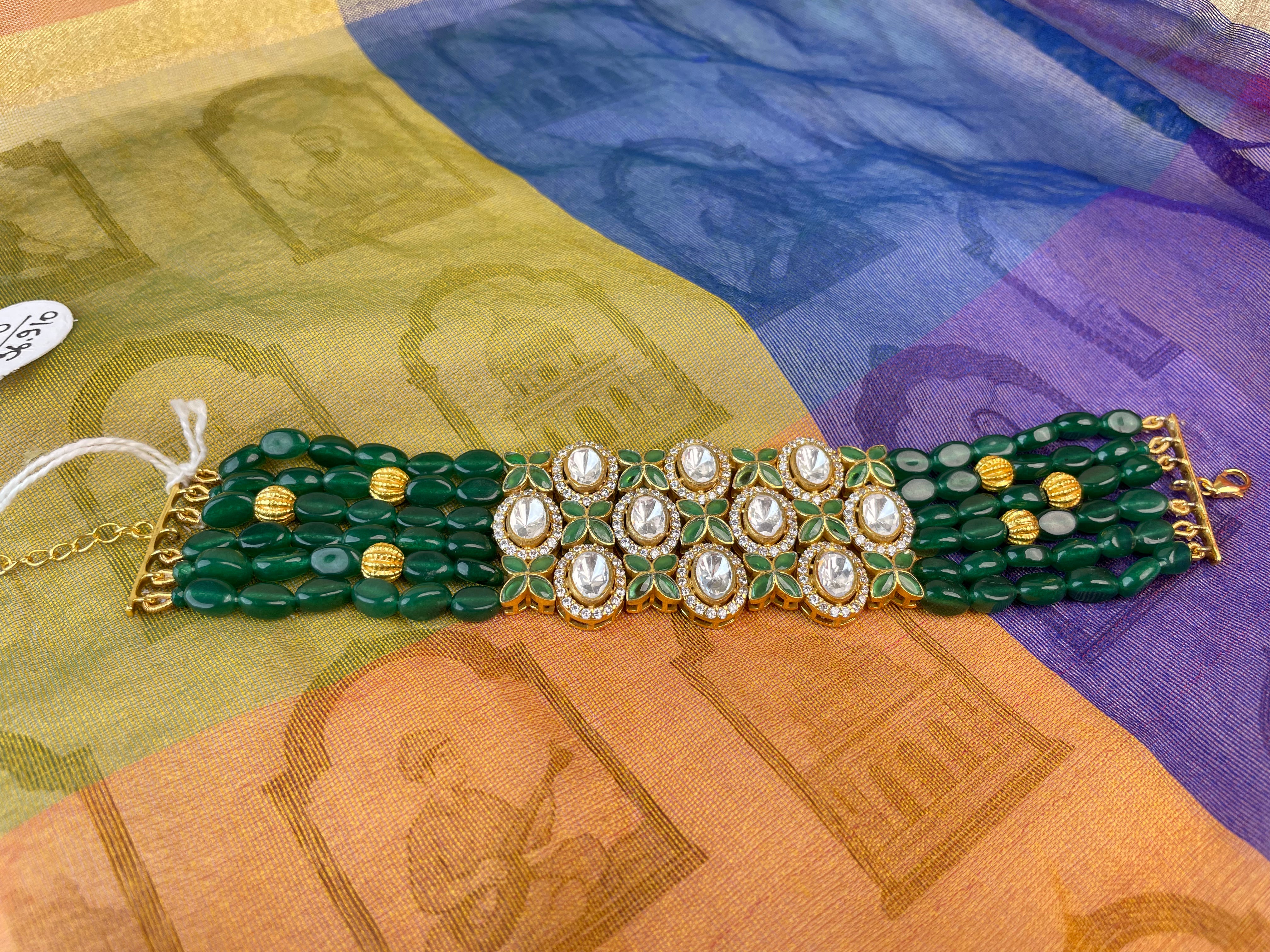 A green beaded bracelet with gold-plated details and central white stone embellishments, displayed on a multicolored patterned fabric background.