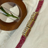 A close-up of a traditional Indian jewelry piece featuring a deep red beaded strap with three ornate gold-toned decorative beads embellished with white pearls and red inlay, displayed on a cream-colored fabric next to a wooden ring holder.