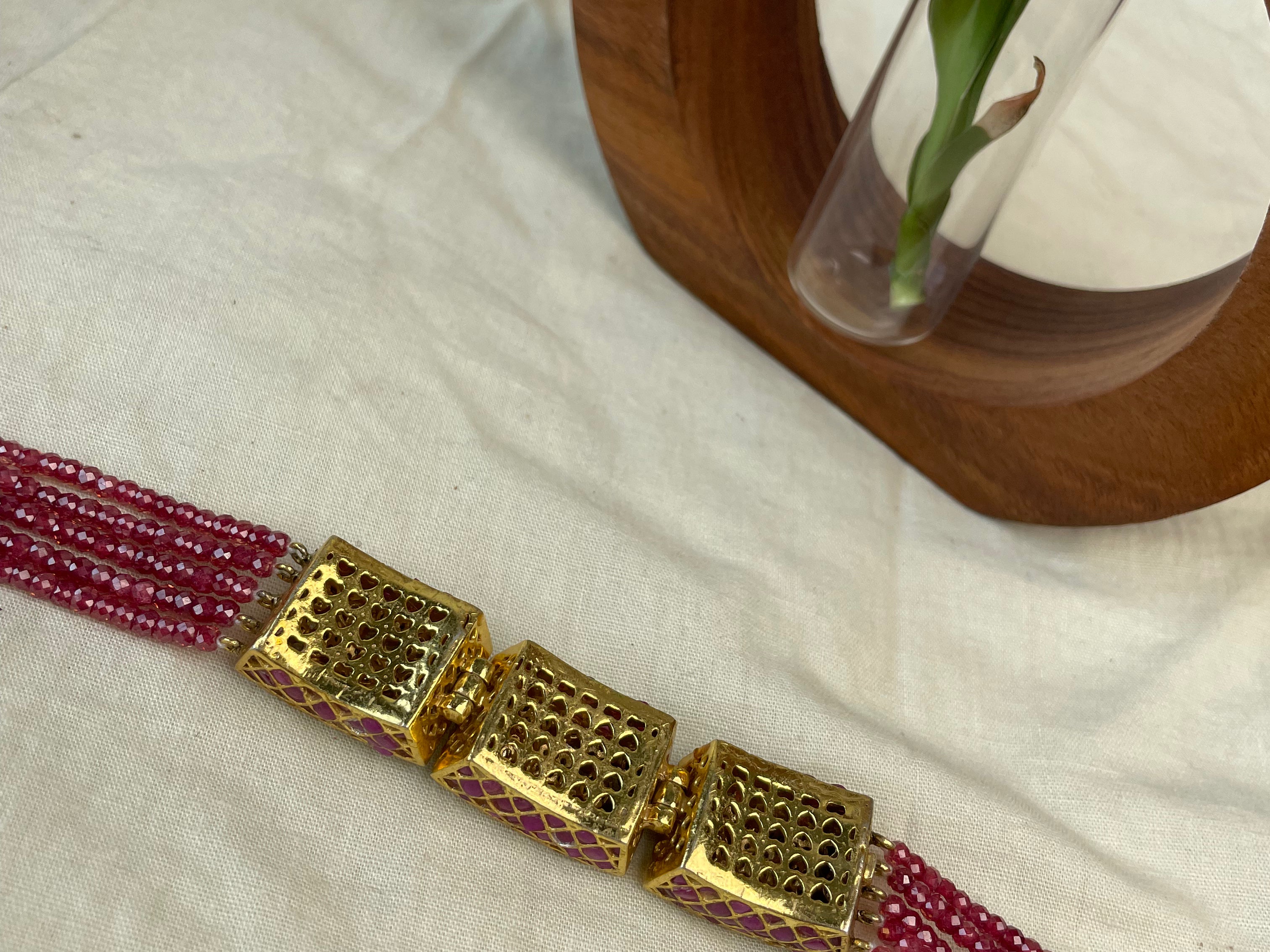 A beaded bracelet with multiple strands of deep red beads and three large textured gold-toned square links, displayed on a cream fabric surface with a wooden ring holder in the background.