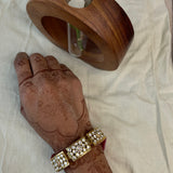 A close-up of a person's hand with henna wearing a gold-toned traditional Indian bracelet adorned with white stones, next to a wooden ring holder on a white fabric background.