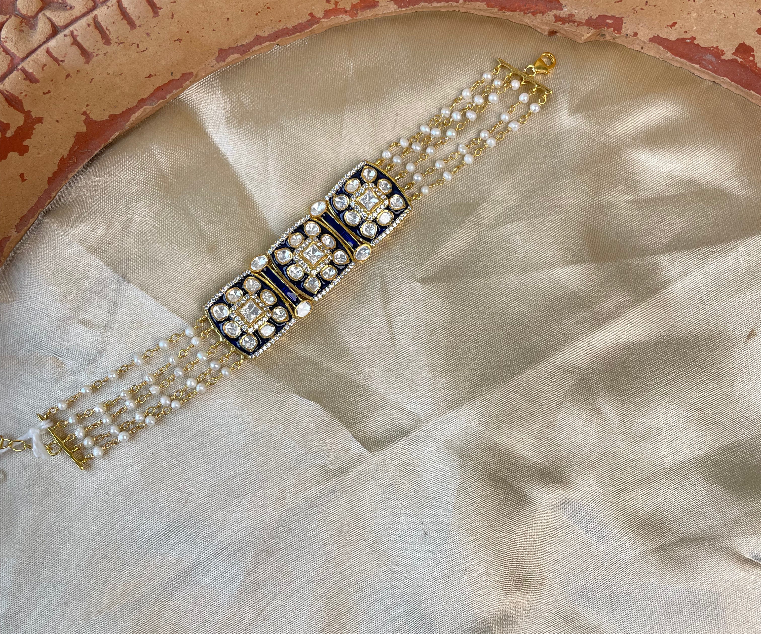 A traditional Indian Kundan bracelet with a central rectangular gold-plated centerpiece adorned with black enamel and white stones, flanked by triple rows of white pearls on a beige satin fabric with a terracotta plate prop.