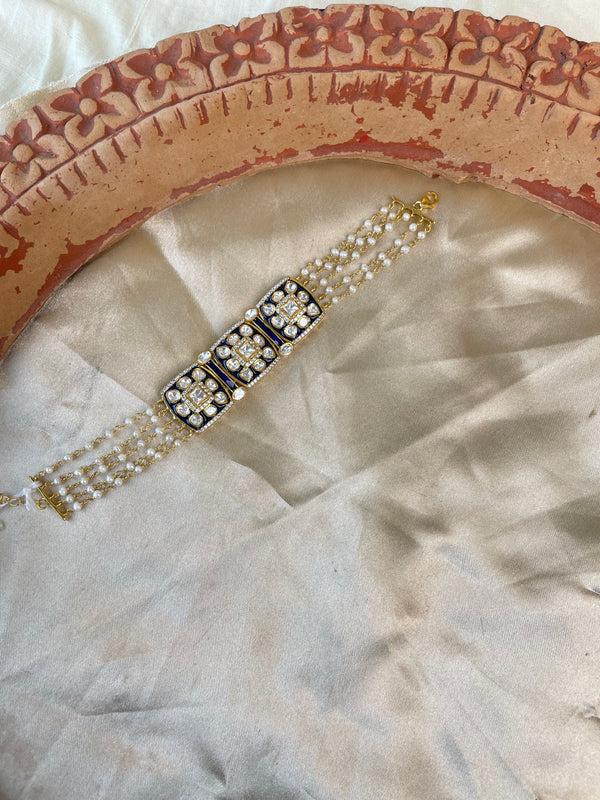 A traditional Indian Kundan bracelet with a central rectangular gold-plated centerpiece adorned with black enamel and white stones, flanked by triple rows of white pearls on a beige satin fabric with a terracotta plate prop.