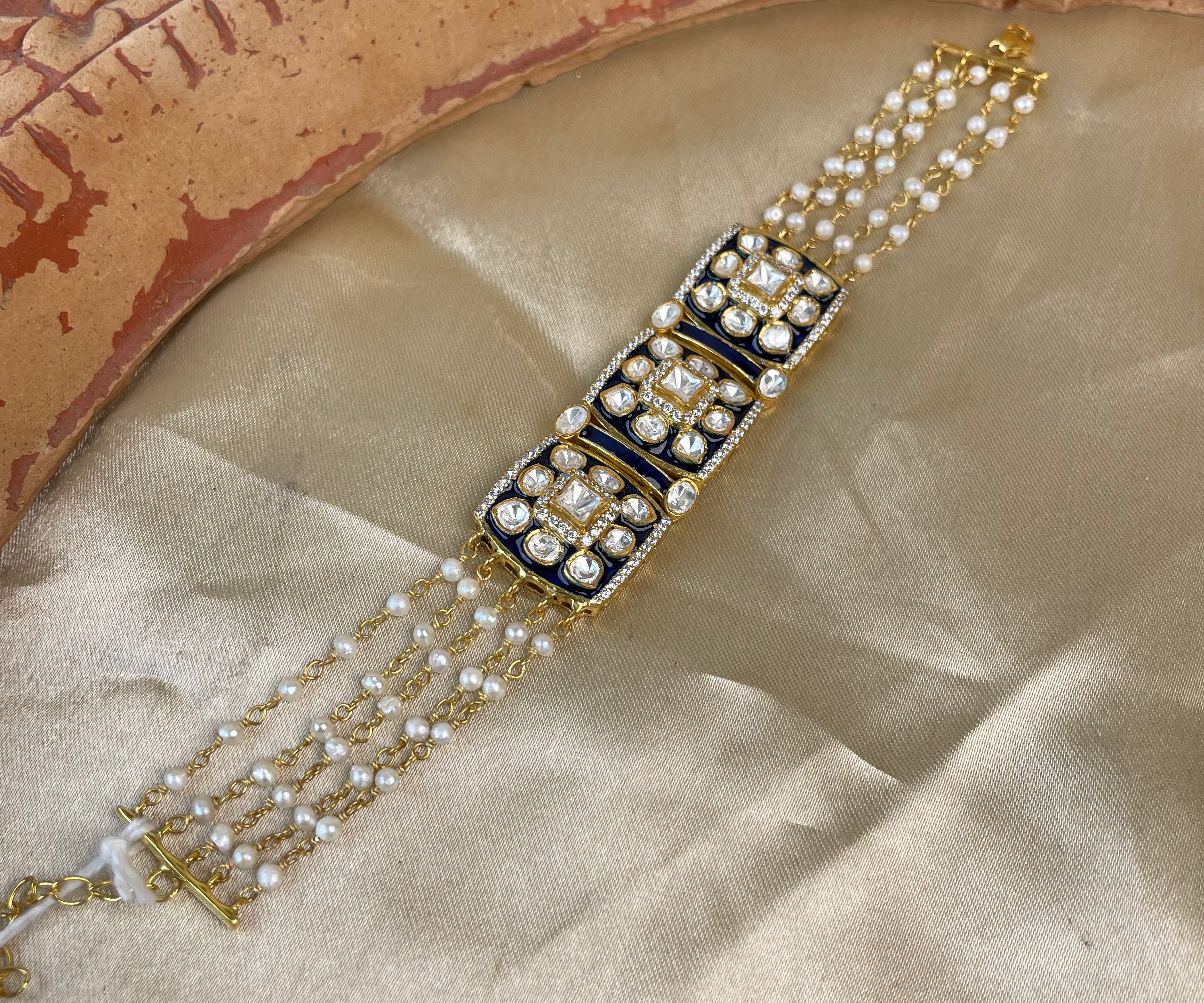 A gold-plated traditional Indian Kundan bracelet with black enamel panels and white stones, displayed on a beige satin fabric next to a carved wooden bowl.