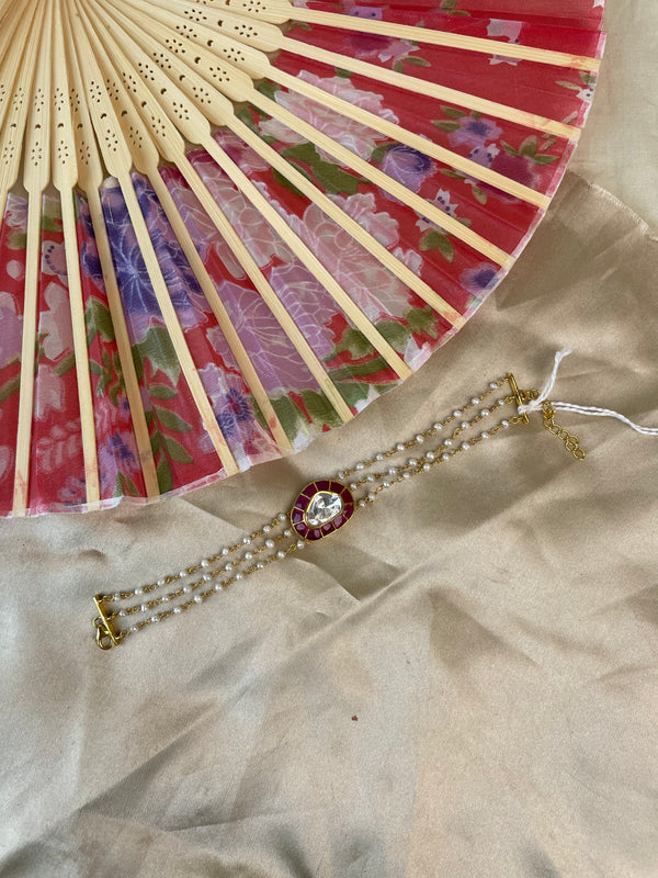 A gold-toned multi-strand pearl bracelet with a central red and gold floral medallion, displayed on beige satin fabric next to a red and beige folding fan.