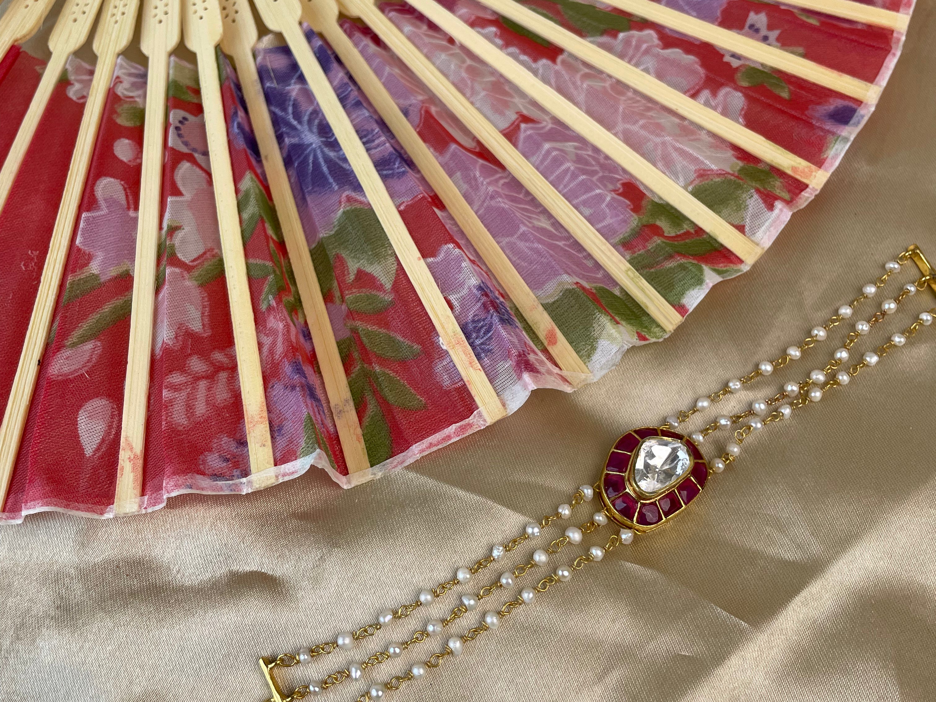 A gold-toned multi-strand pearl bracelet with a red and white floral medallion, displayed on beige satin fabric next to a folding fan with a red and purple floral pattern.