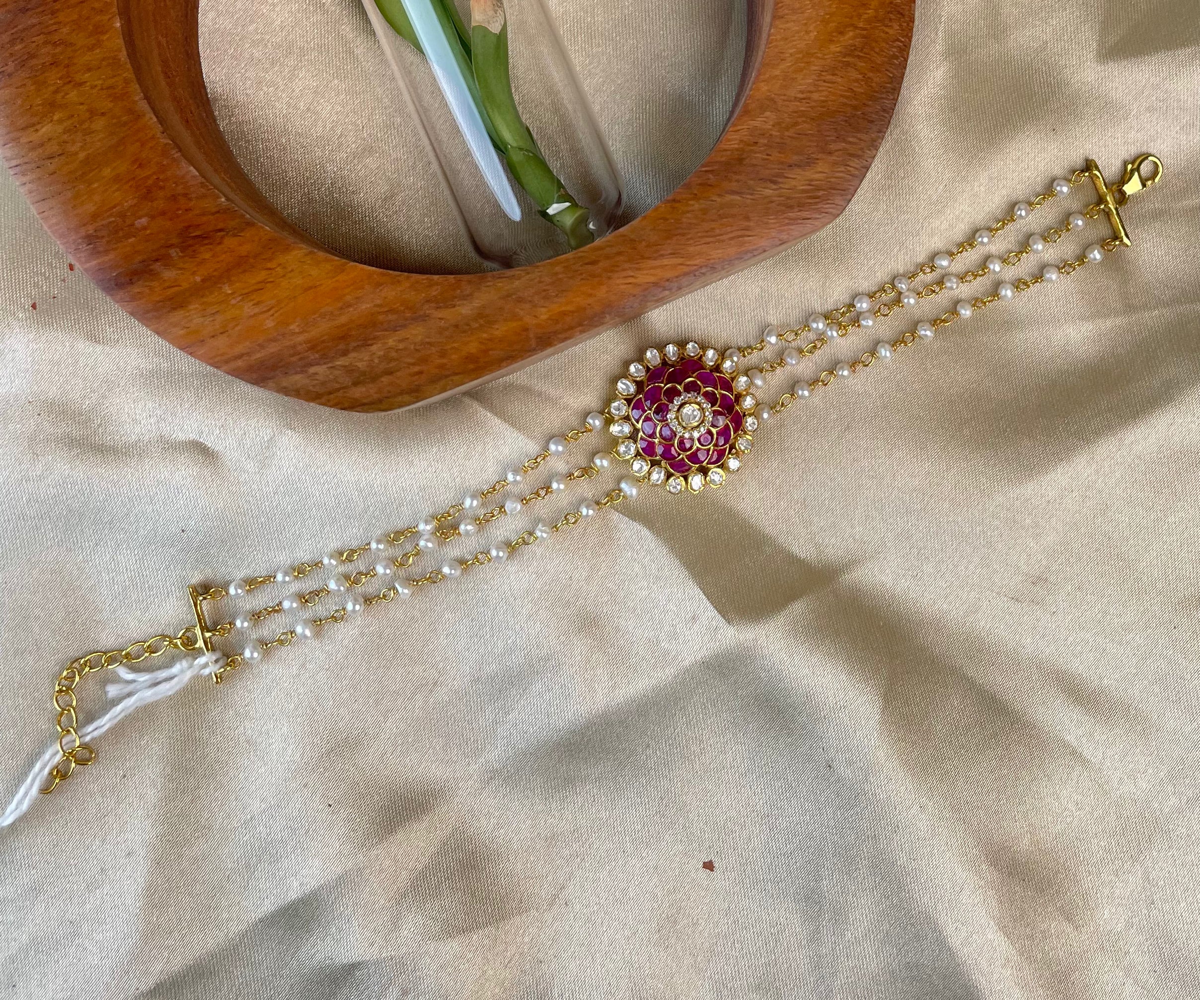 A gold-plated beaded bracelet with a central red and gold floral pendant, displayed on a beige satin fabric next to a wooden ring holder.