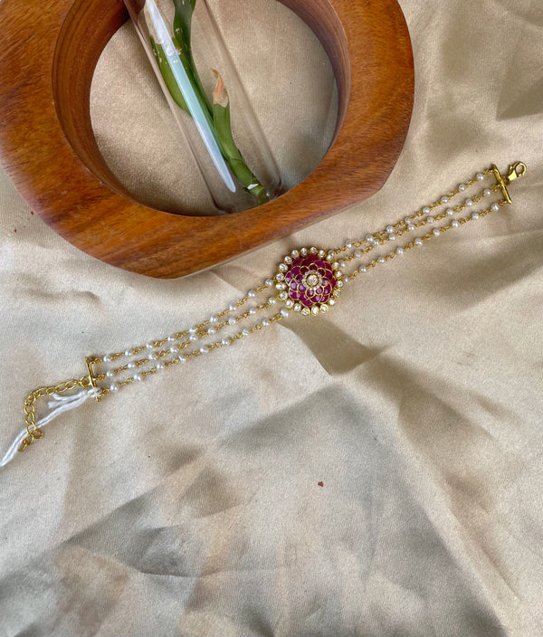 A gold-plated beaded bracelet with a central red and gold floral pendant, displayed on a beige satin fabric next to a wooden ring holder.
