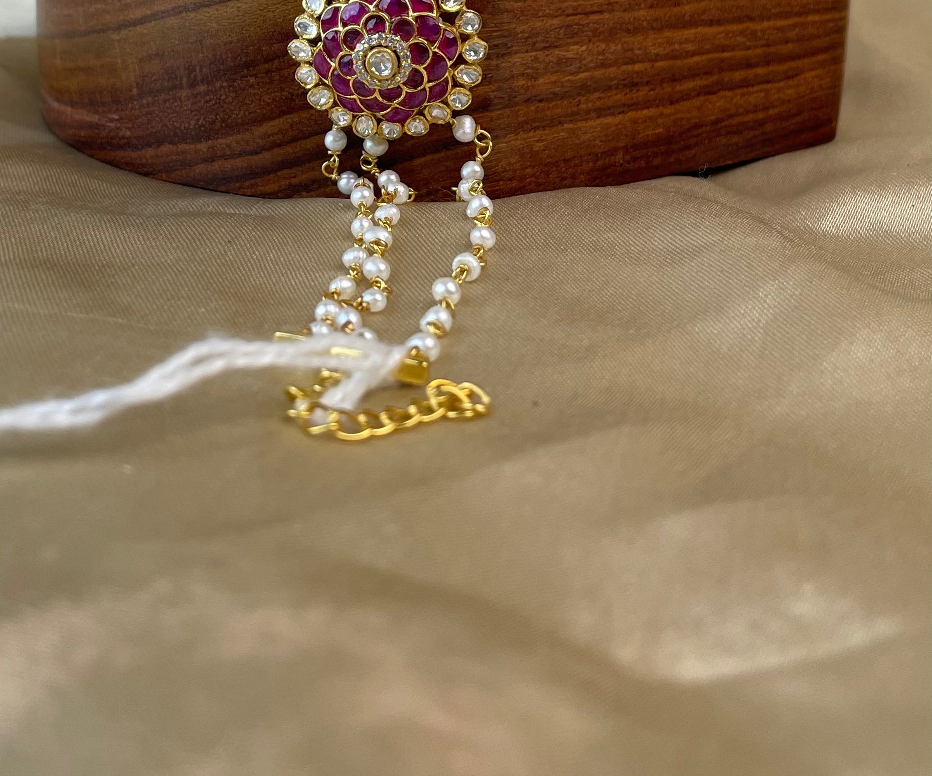 A gold and pink floral bracelet with white pearls and a central clear stone, draped over a wooden stand on a beige satin fabric background.