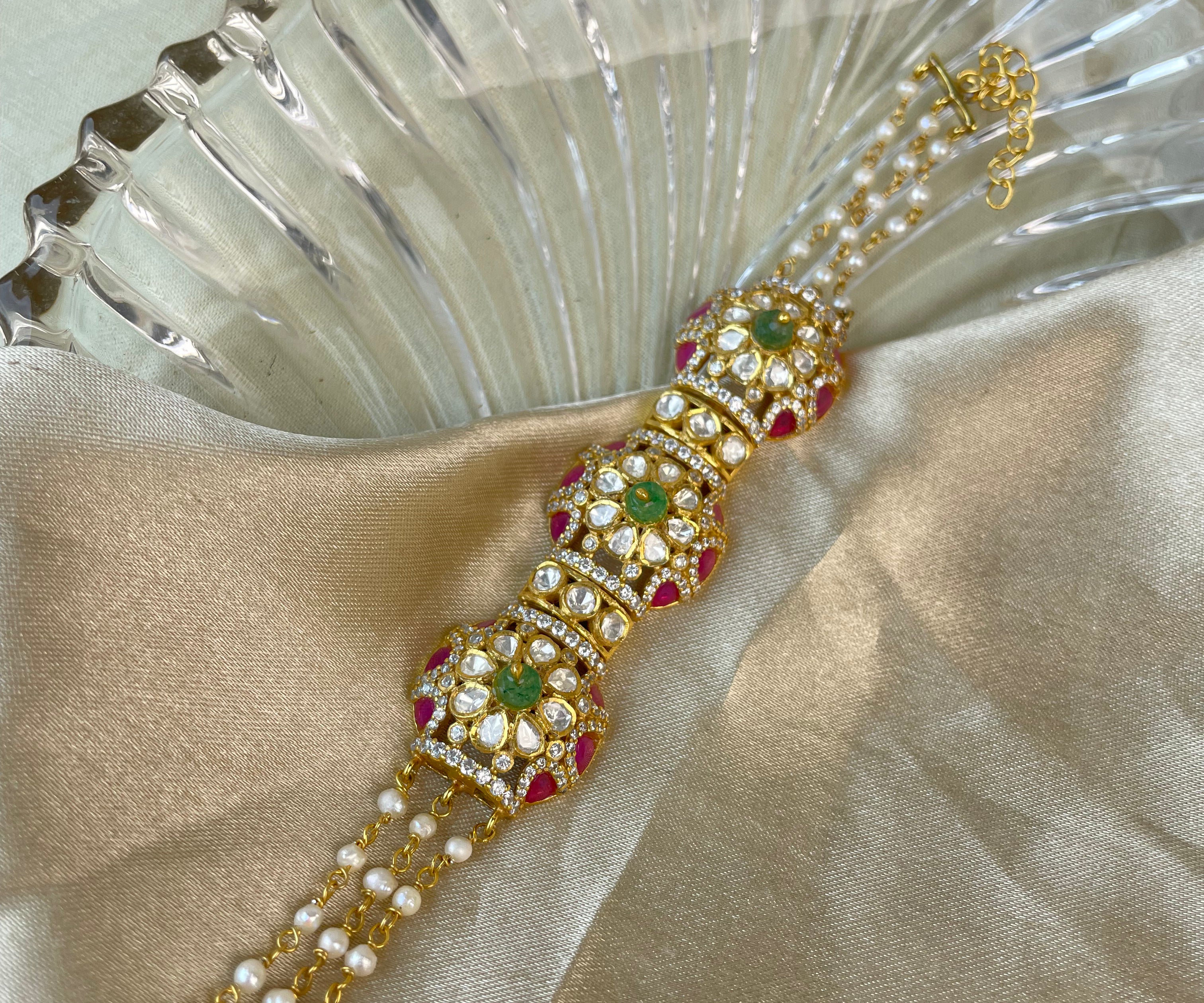 A gold-plated traditional Indian Kundan bracelet adorned with white pearls, green stones, and red stones, displayed on a beige satin fabric next to a clear glass bowl.