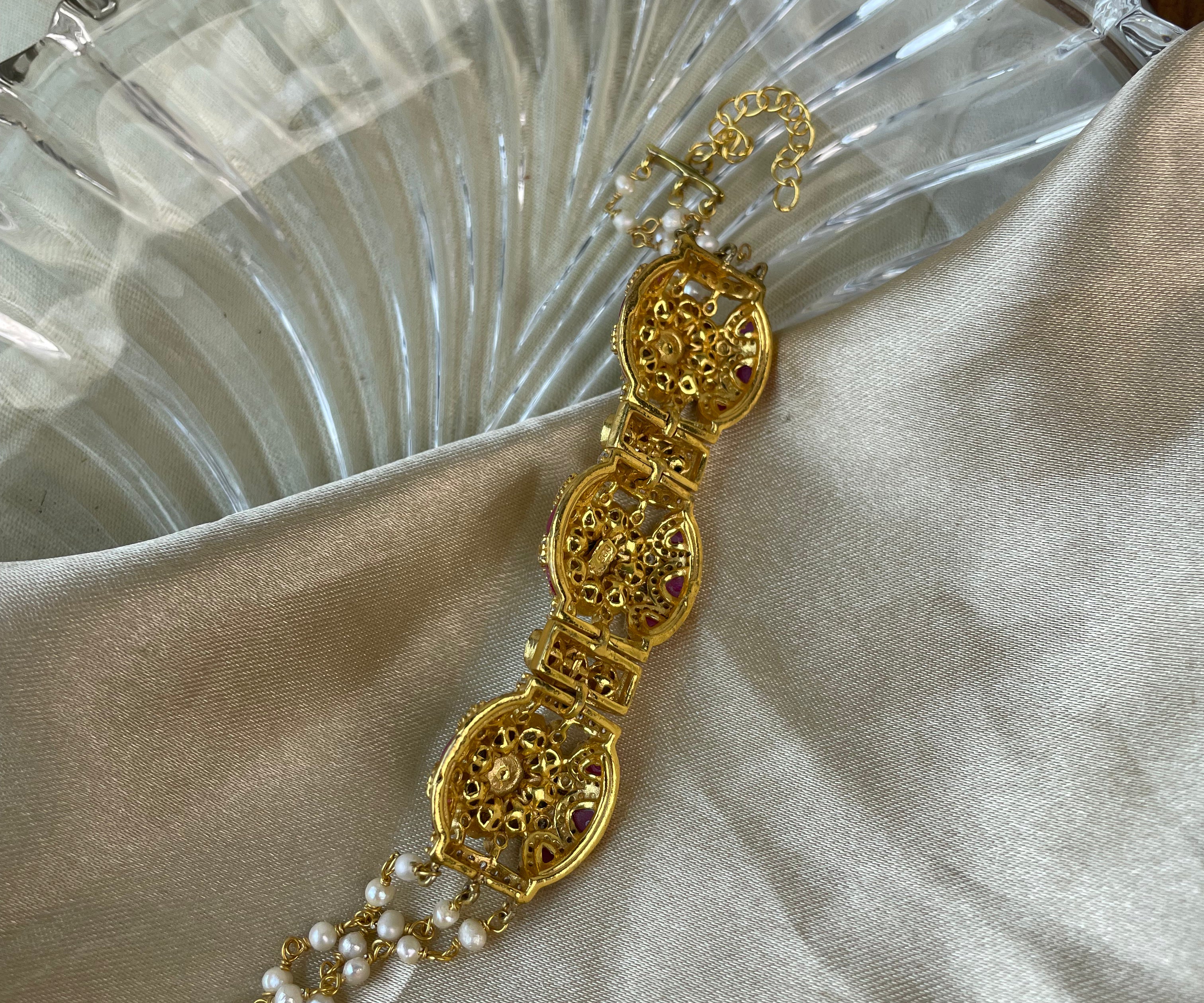 A gold-toned ornate bracelet with oval filigree motifs and pearl strands, displayed on draped satin fabric next to a clear ribbed glass dish.