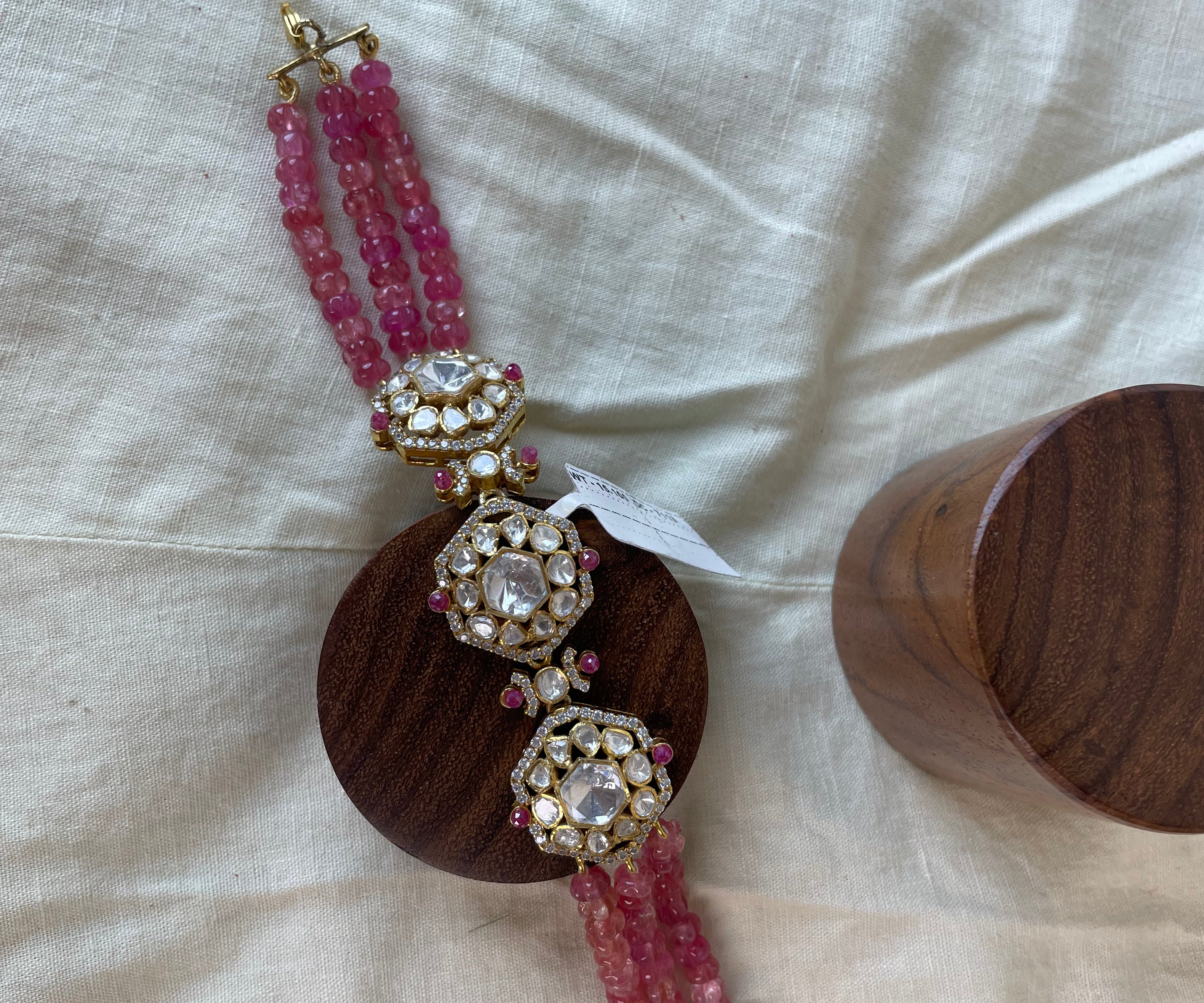 A traditional Indian bridal bracelet with pink gemstone beads, ornate gold-plated filigree details, and clear stones, displayed on a wooden stand next to a wooden box on a beige fabric background.