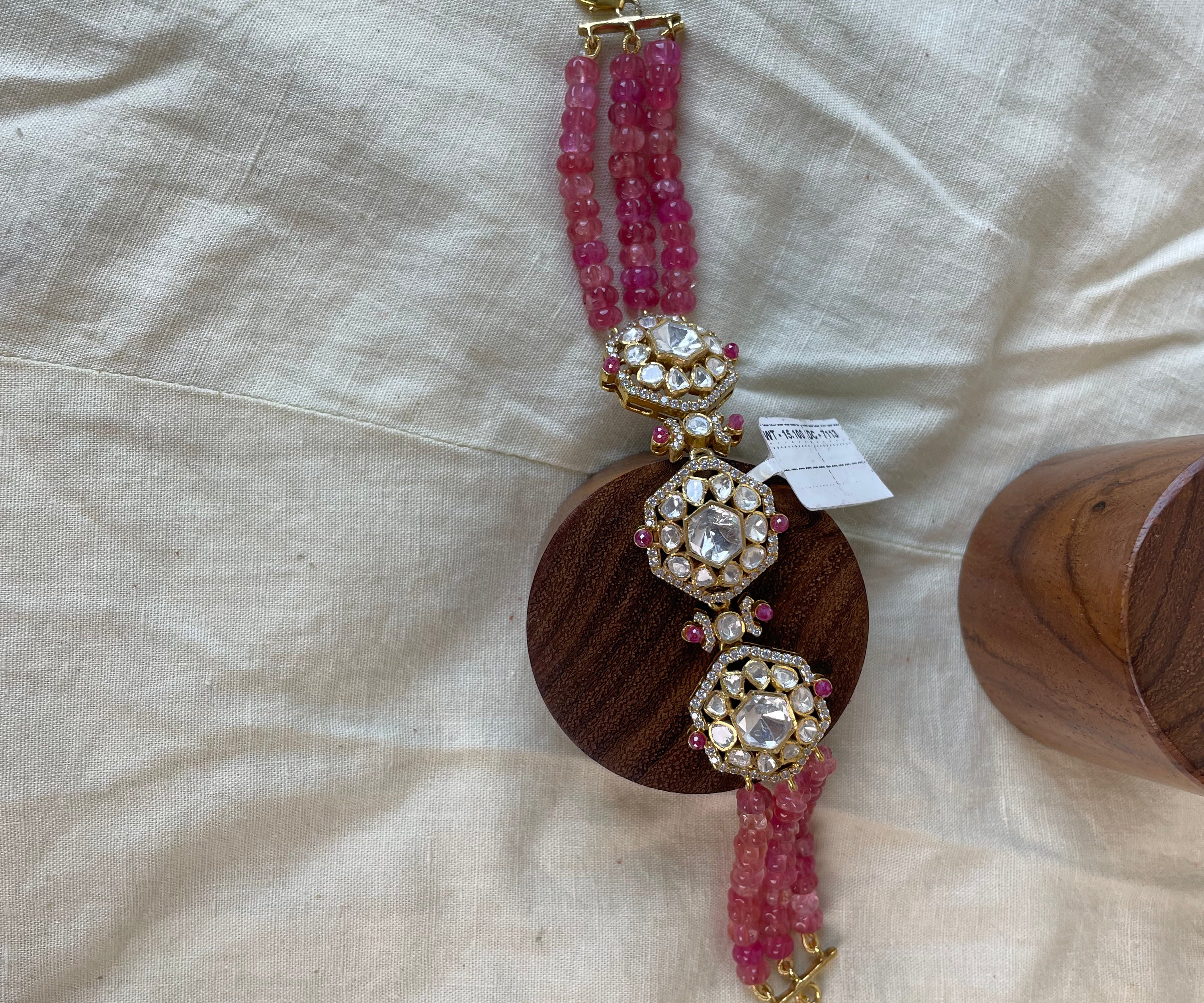 A pink gemstone beaded bracelet with ornate gold-toned filigree and clear crystal embellishments, displayed on a round wooden stand over a beige fabric background.