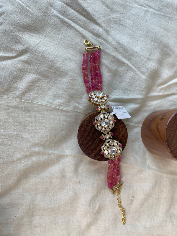 A pink gemstone beaded bracelet with ornate gold-toned filigree and clear crystal embellishments, displayed on a round wooden stand over a beige fabric background.