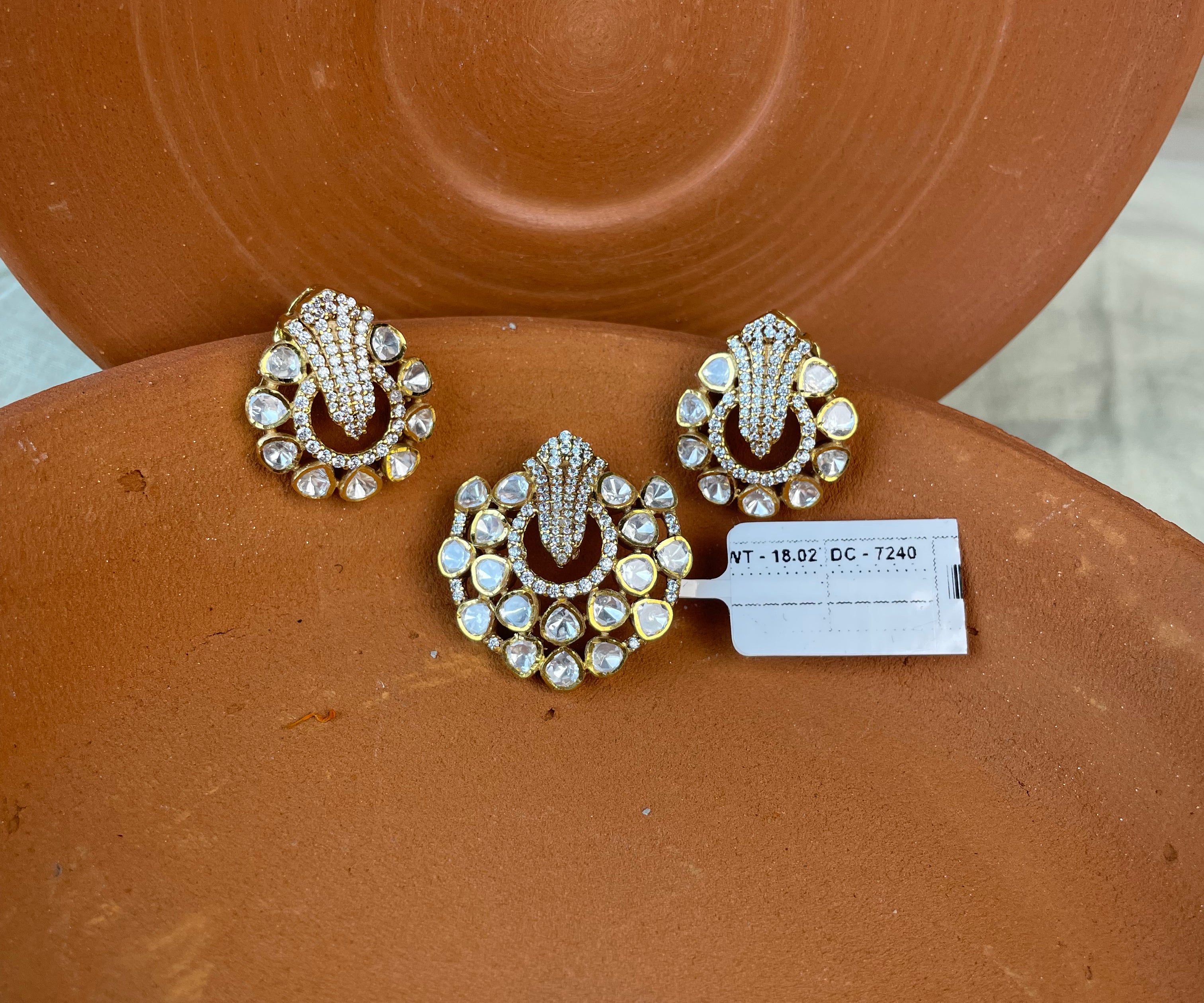 Three ornate gold-toned pendant and earrings with clear rhinestones and dark red enamel details, arranged on a terracotta-colored ceramic surface with a small white inventory tag.