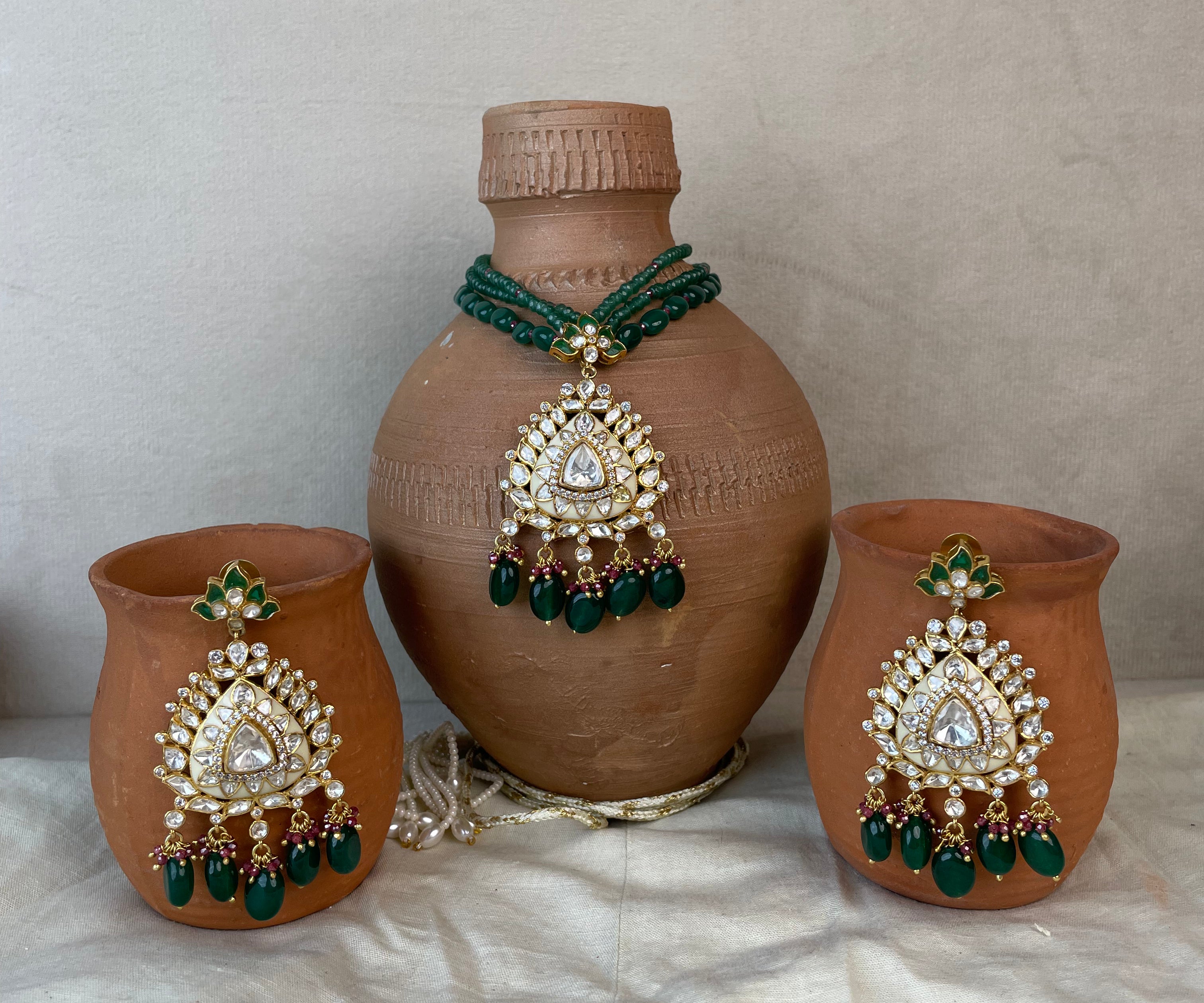 Three terracotta clay vessels decorated with ornate gold and green jewelry-style embellishments, including a large vase in the center and two matching cups on either side, arranged on a draped white fabric surface against a neutral wall.