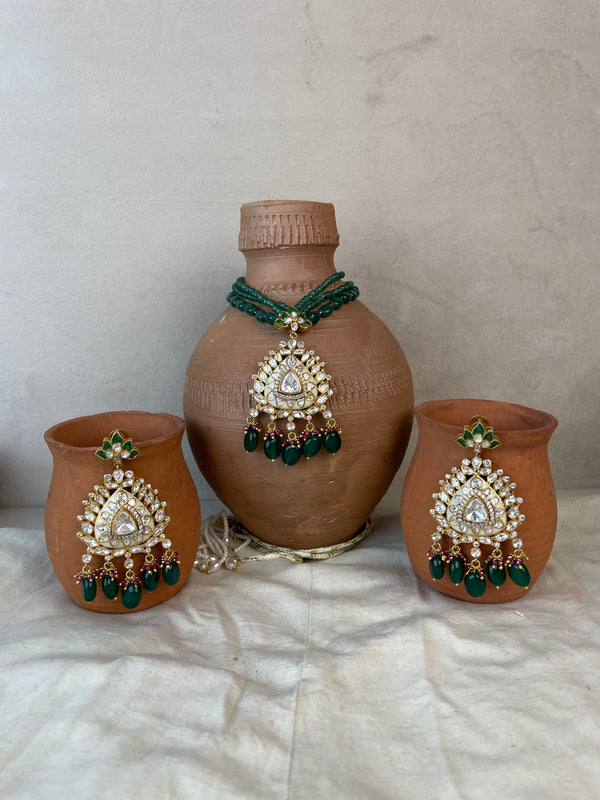 Three terracotta clay vessels decorated with ornate gold and green jewelry-style embellishments, including a large vase in the center and two matching cups on either side, arranged on a draped white fabric surface against a neutral wall.