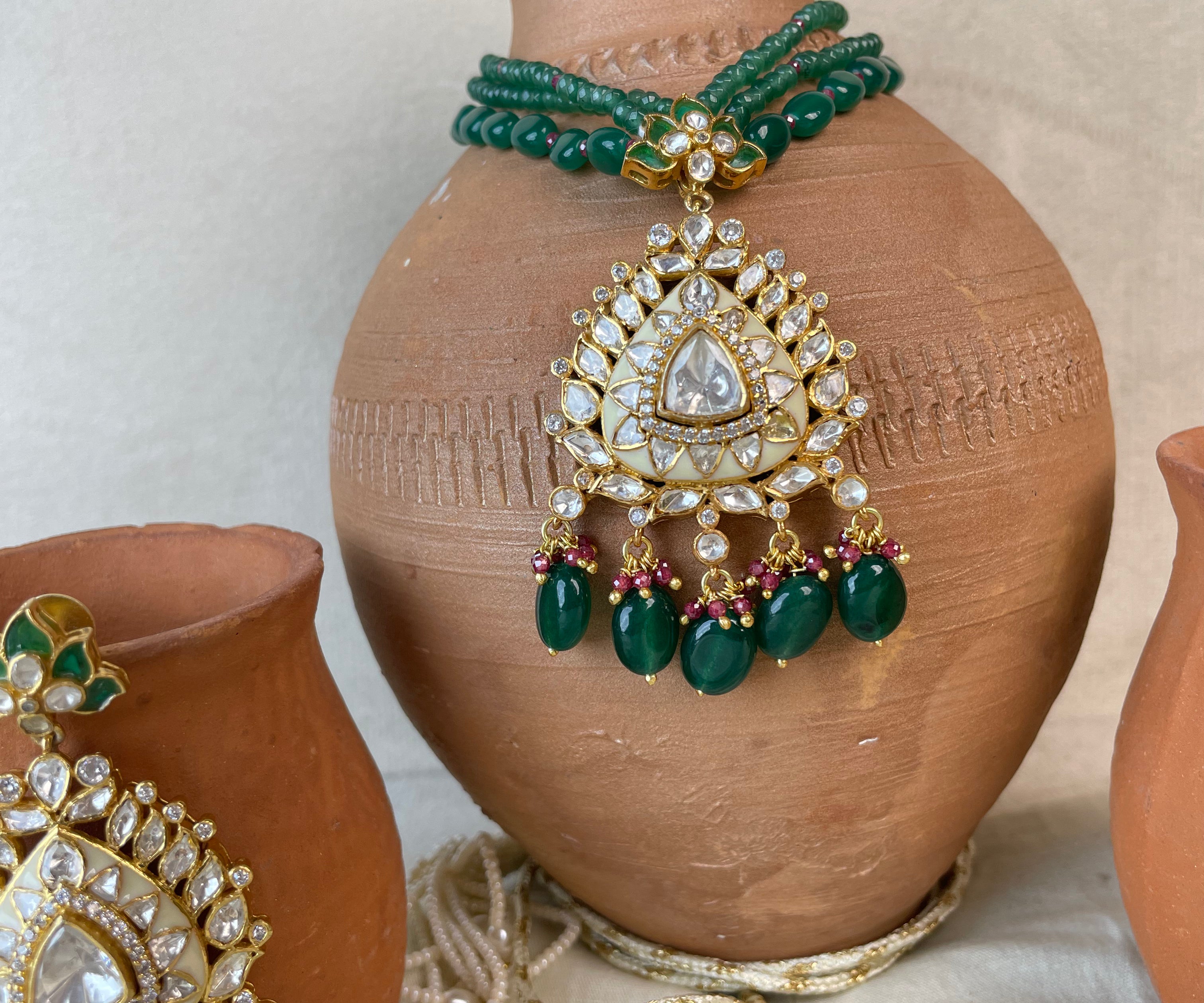 Gold and green beaded jewelry displayed on terracotta pots against a neutral fabric backdrop. The set includes a necklace with a large teardrop-shaped gold pendant set with clear stones and green beads, a matching headpiece, and coordinating green beaded bracelets.
