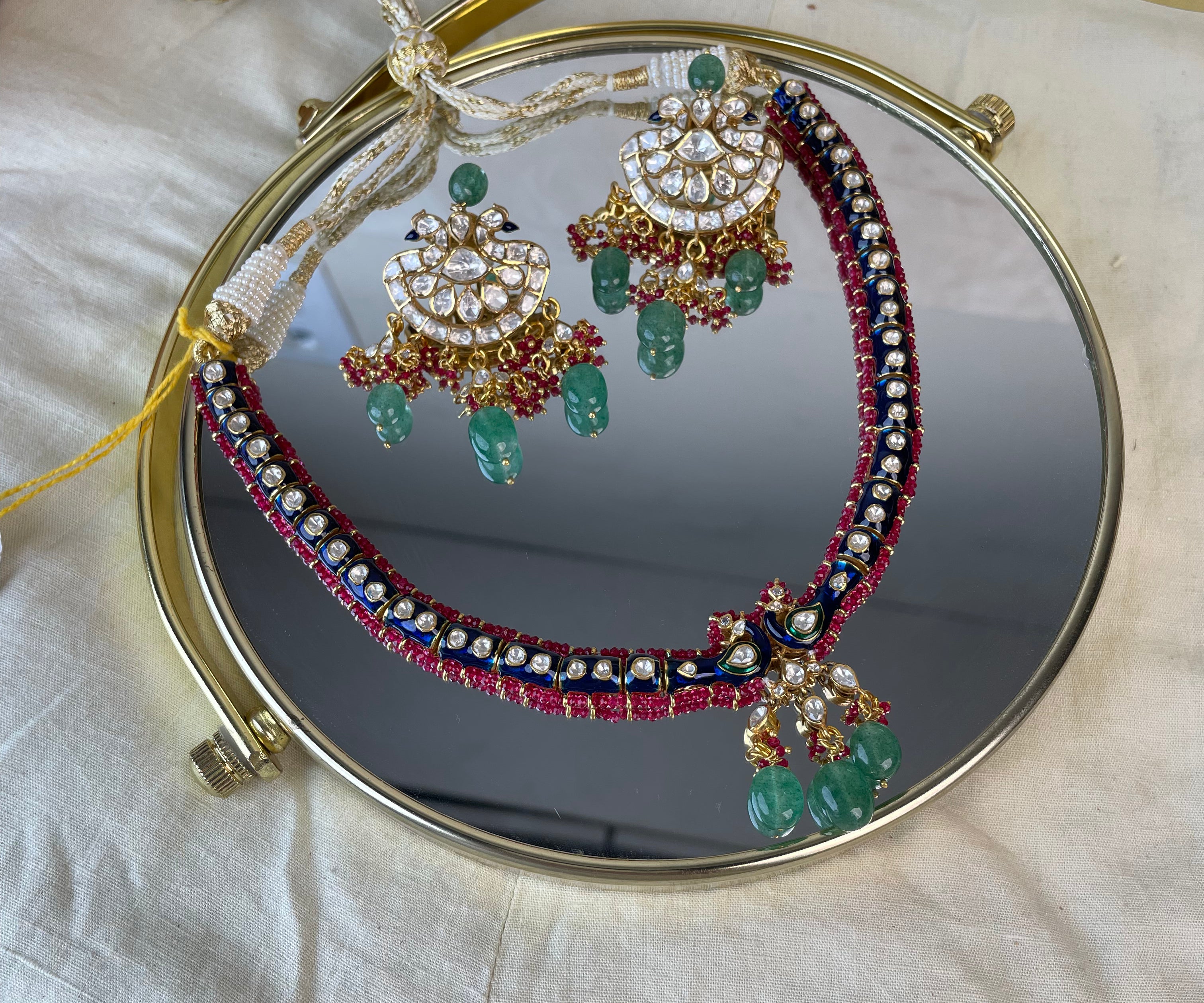 A traditional Indian jewelry set displayed on a round gold-framed mirror over a white fabric surface. The set includes a necklace and matching earrings with gold-plated metalwork, white stones, and green beadwork and hanging green beads.