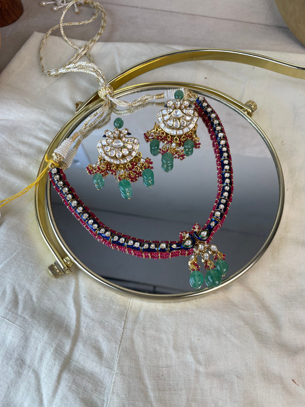 A traditional Indian jewelry set displayed on a round gold-framed mirror over a white fabric surface. The set includes a necklace and matching earrings with gold-plated metalwork, white stones, and green beadwork and hanging green beads.