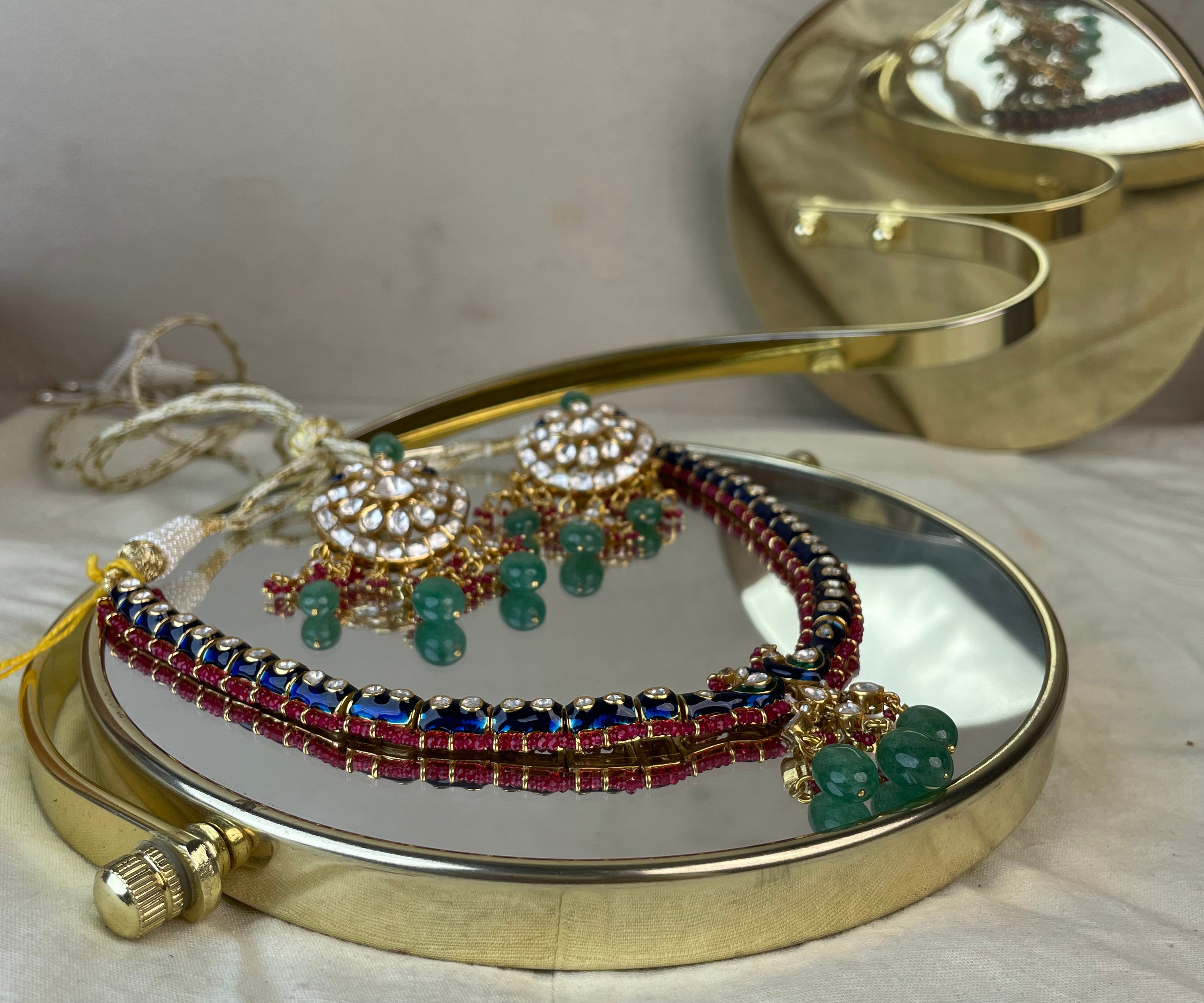 A gold-plated traditional Indian necklace and matching earrings displayed on a round mirror, featuring red and blue beads, white stones, and green bead drops.