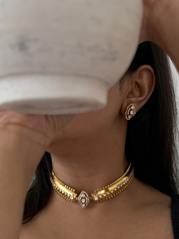A close-up lifestyle shot of a model wearing a gold choker necklace and matching stud earring. The jewelry features gold-plated metal with clear pavé-set stones, including a central teardrop-shaped stone.