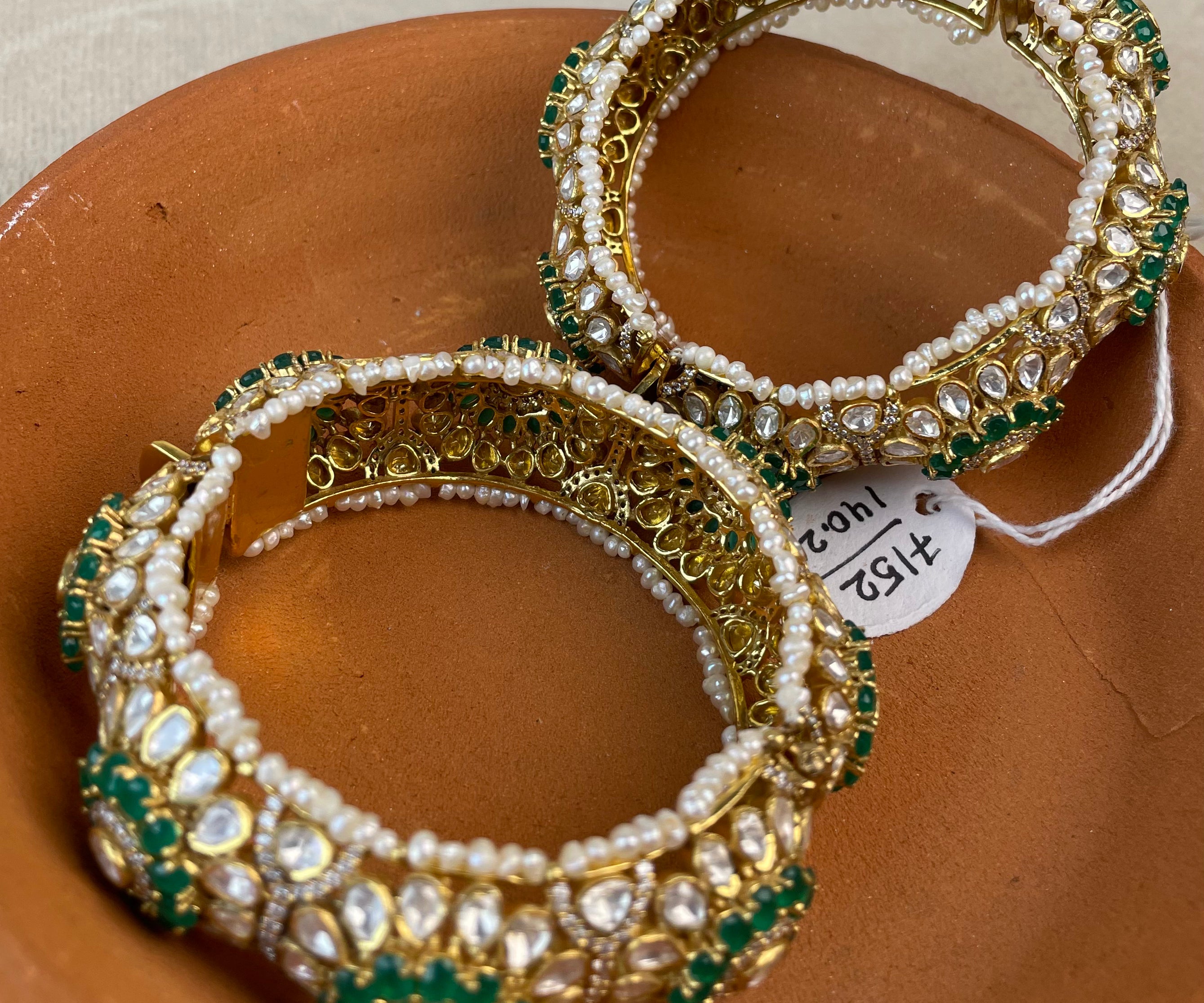 Two ornate bangles decorated with white pearls and green stones, displayed on a terracotta plate.